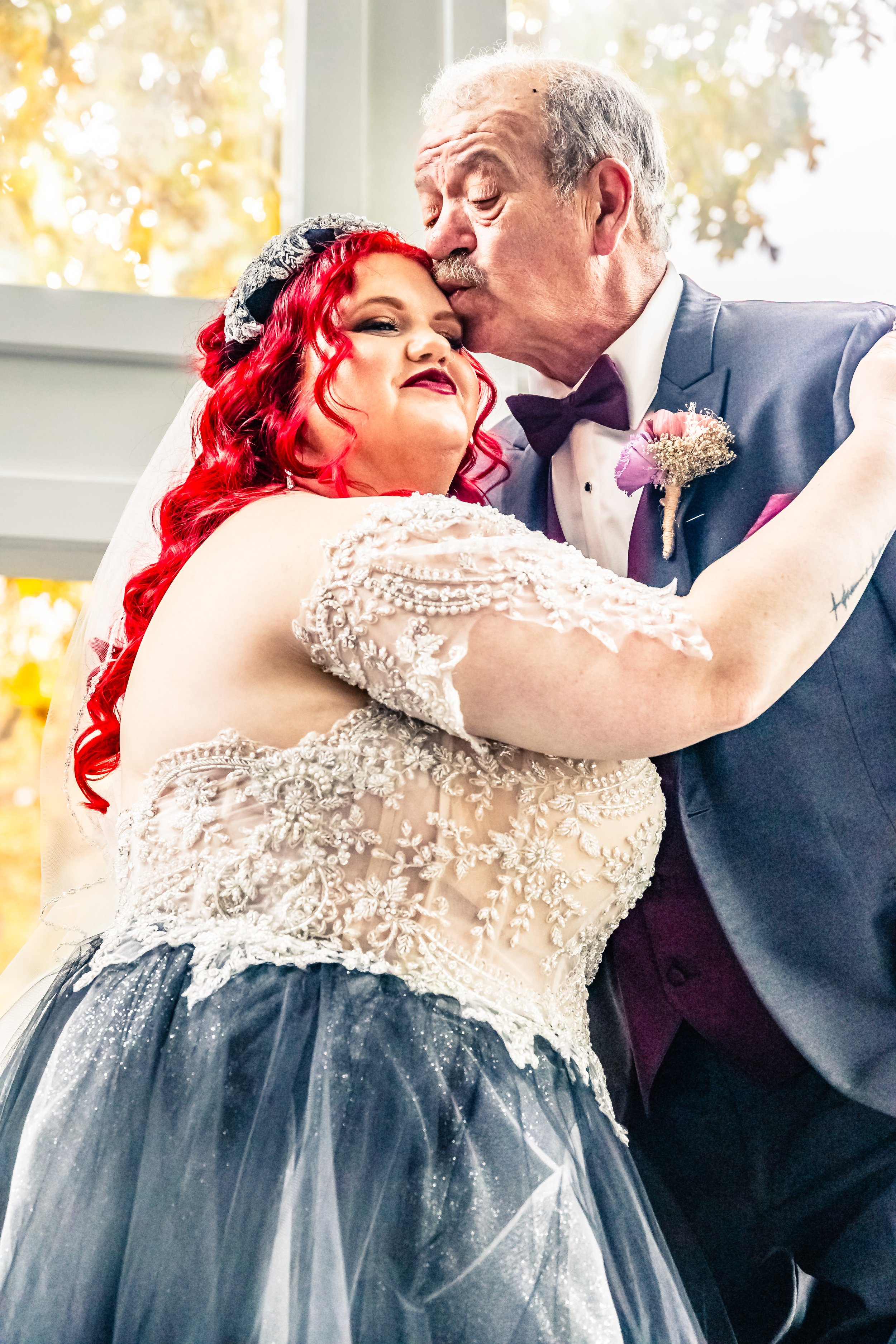 An elderly man with gray hair and a mustache kissing a woman with bright red hair on the forehead at a wedding ceremony.