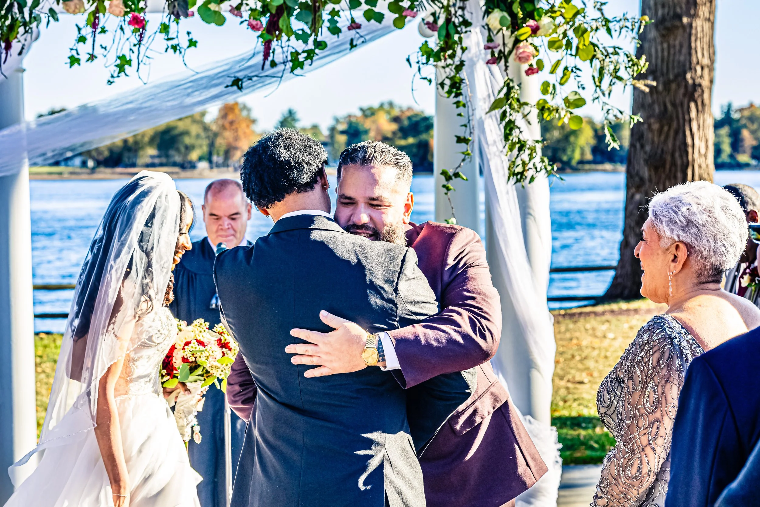 A wedding ceremony outdoors by a body of water, with a bride, groom, and an officiant, as two men hug in front, surrounded by guests and decorative greenery.