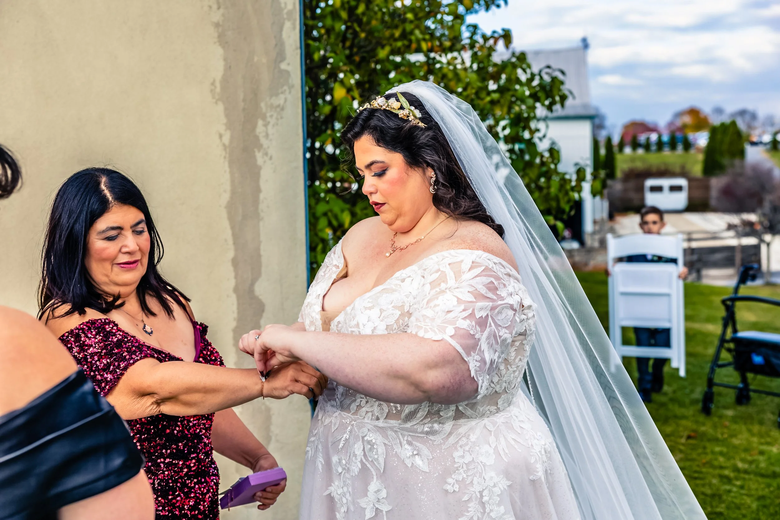 Bride in a lace wedding dress with a veil is being assisted by a woman in a red and black dress during her wedding outside.