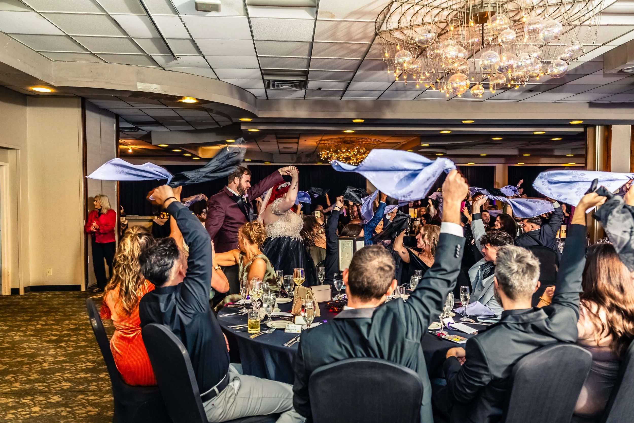 Guests at a wedding reception celebration, with some guests waving blue napkins and a couple dancing in the center of the room.