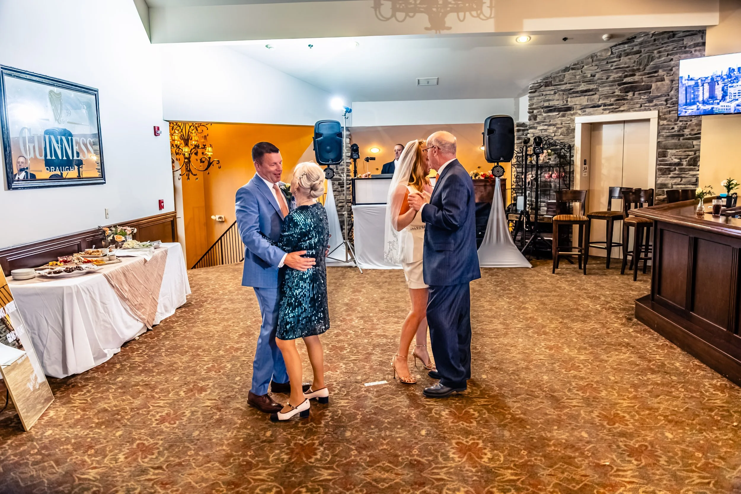 Four people dancing at a wedding reception, with two couples facing each other, in a decorated indoor venue with a buffet table and entertainment equipment in the background.