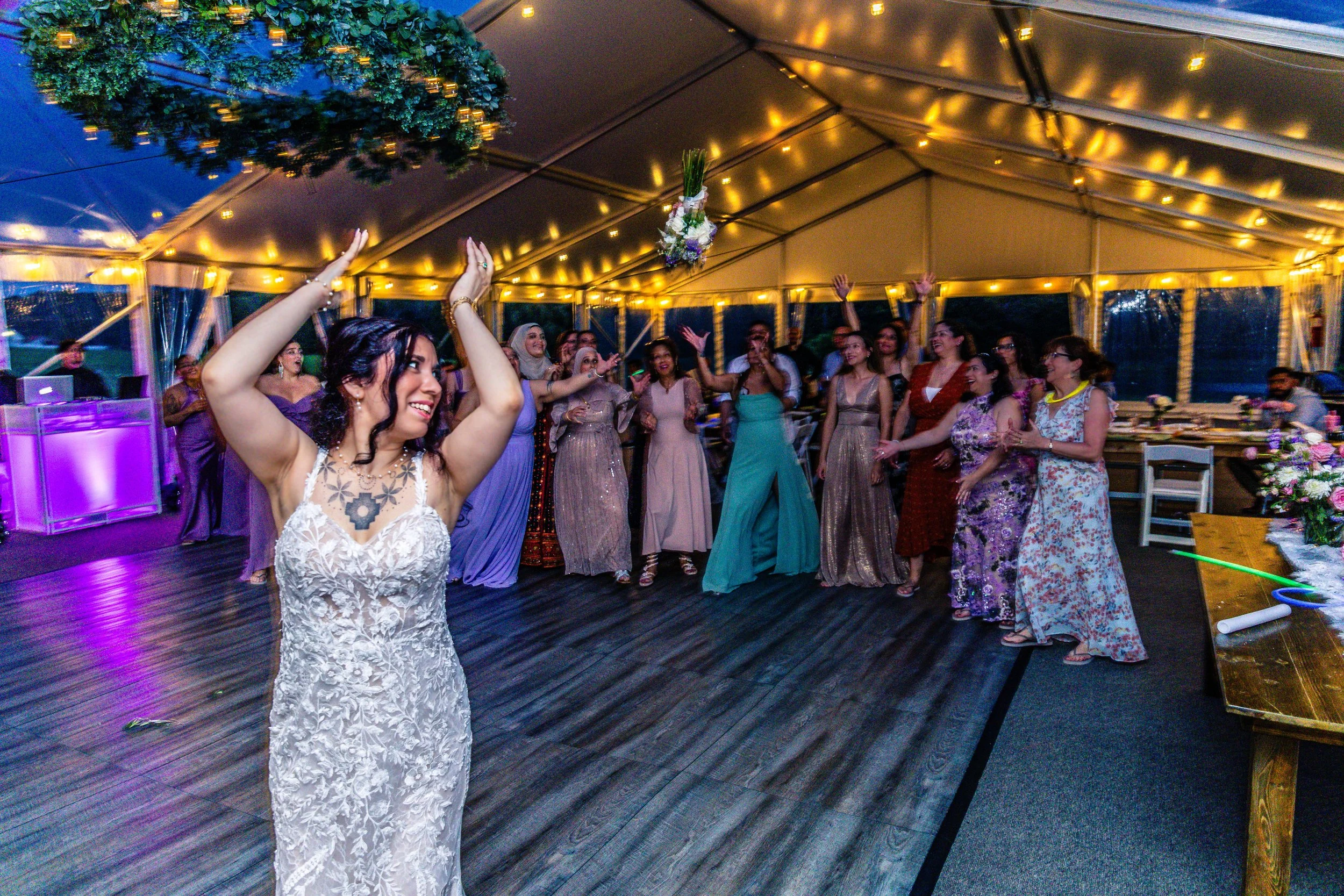 A bride tossing her bouquet to a group of women at a wedding reception under a large tent decorated with string lights.
