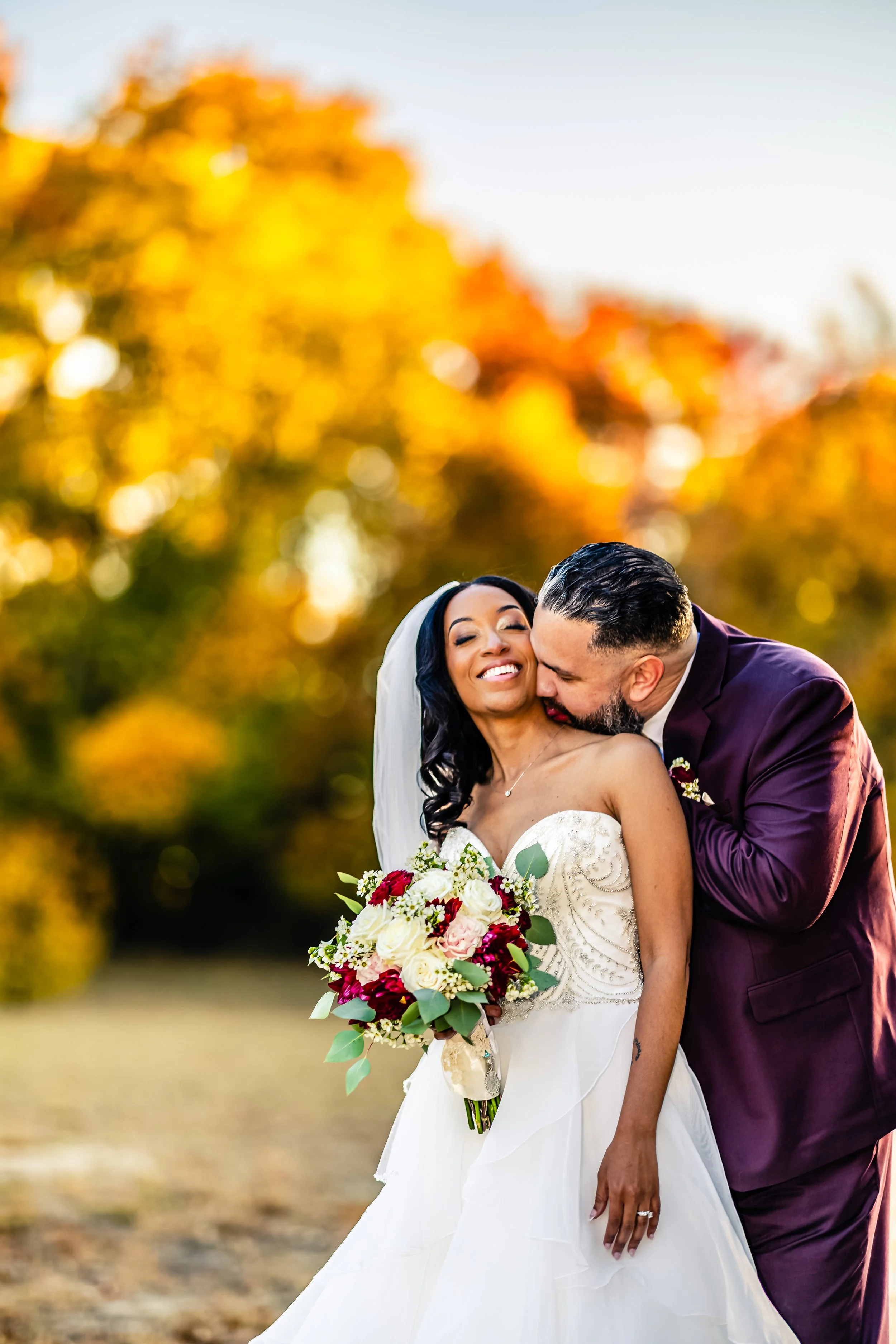 A bride in a white wedding gown holding a bouquet of flowers, smiling as a groom in a dark suit kisses her cheek during an outdoor wedding in autumn.