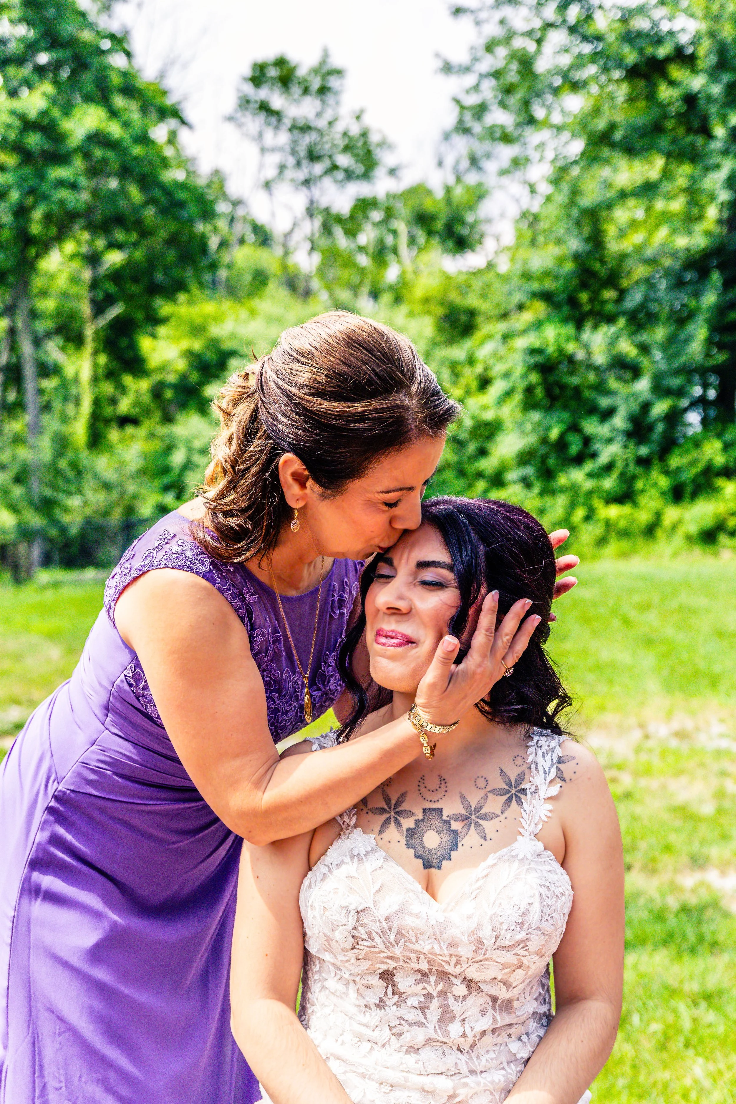 A woman in a purple dress gently cradles a young woman's face as she kisses her forehead, both smiling, outdoors in a green park.