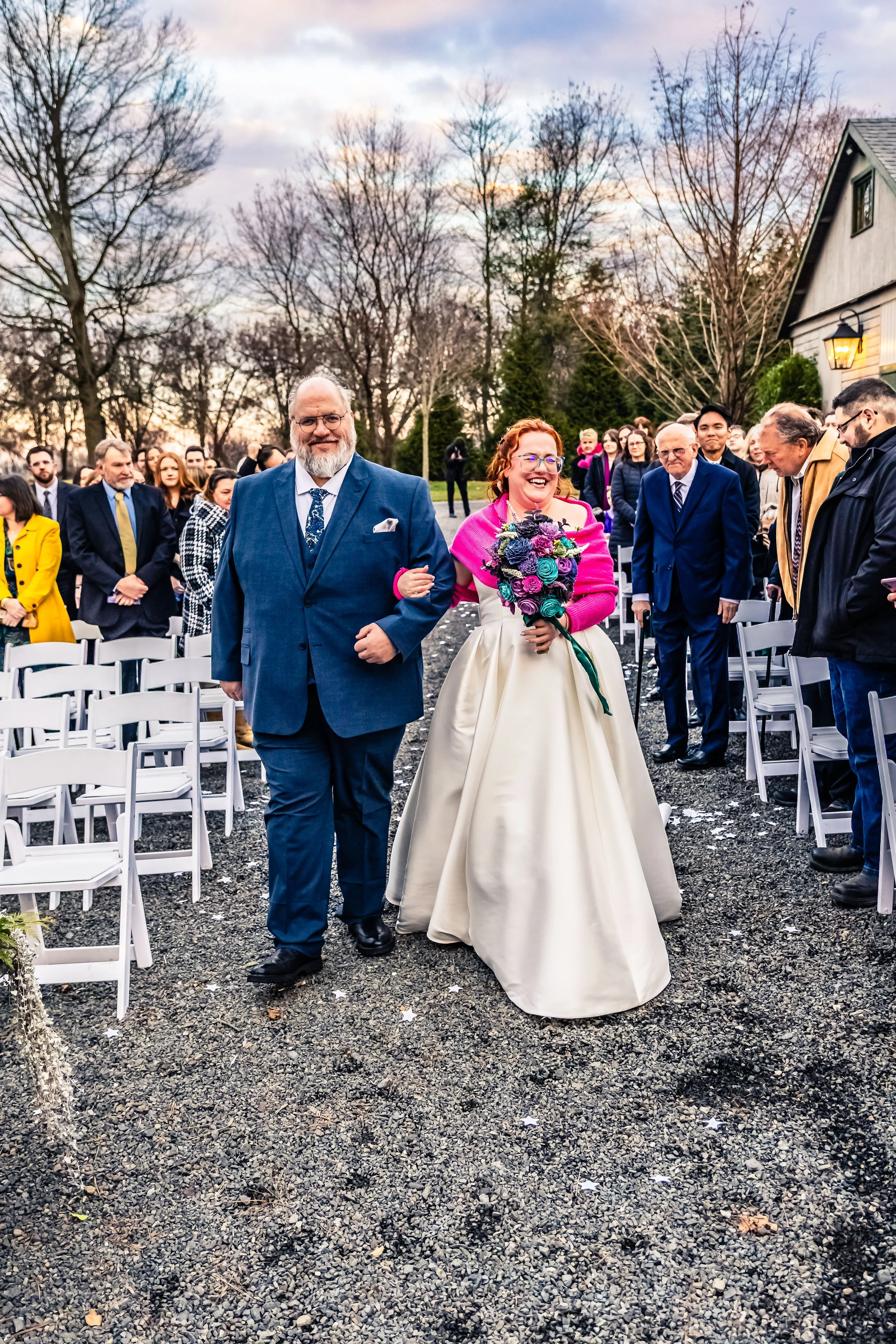 A bride walking down the aisle arm-in-arm with a man in a blue suit, while guests stand and observe outside during a wedding ceremony at dusk. The bride wears a white gown and holds a colorful bouquet, and the man is smiling and dressed formally in a