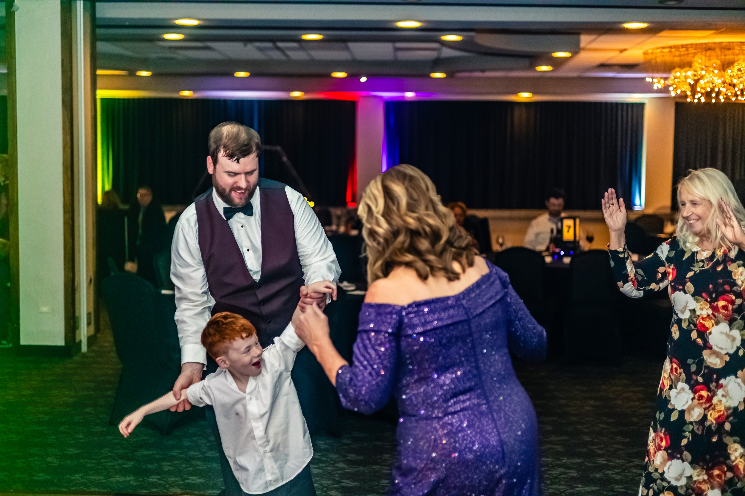 People dancing at a celebration or wedding reception, with colorful lighting and a decorated event space.
