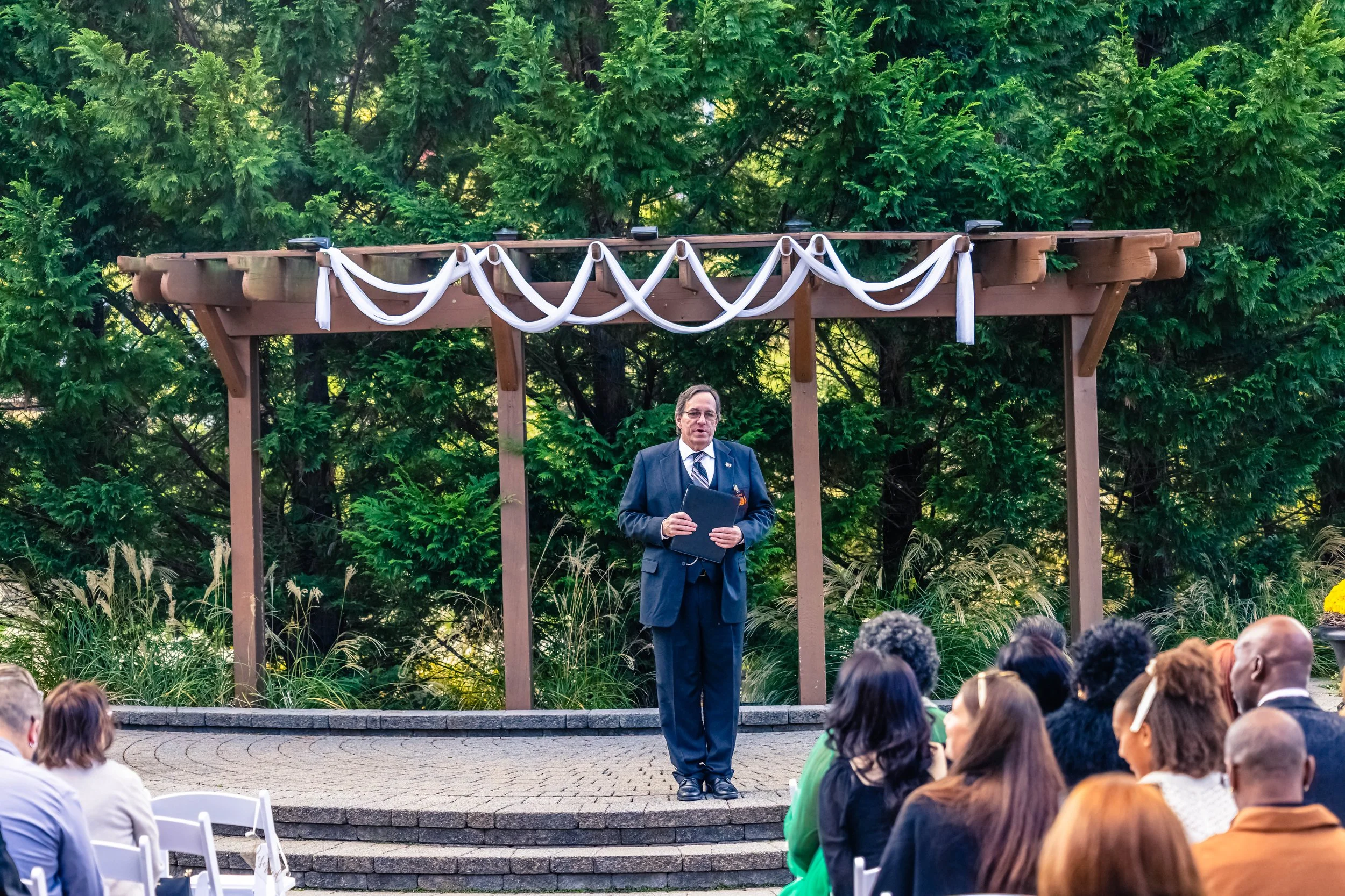 A man in a suit standing on a small outdoor stage speaking to an audience, with a wooden arbor and green trees in the background.