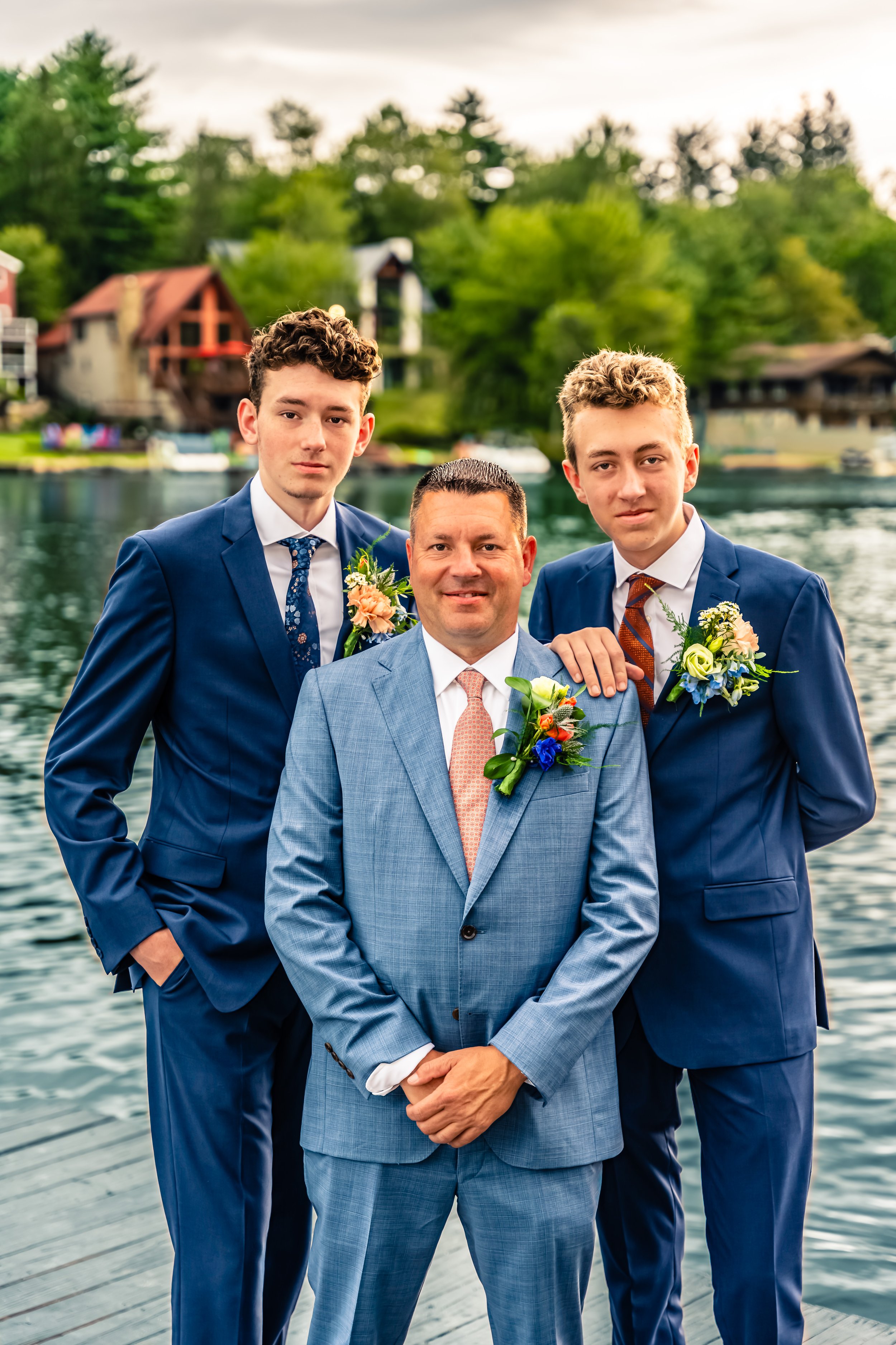 Three men in suits posing by a lake, with trees and houses in the background. The man in the center has a boutonniere, and the other two have boutonnieres and hand on the center man's shoulder.