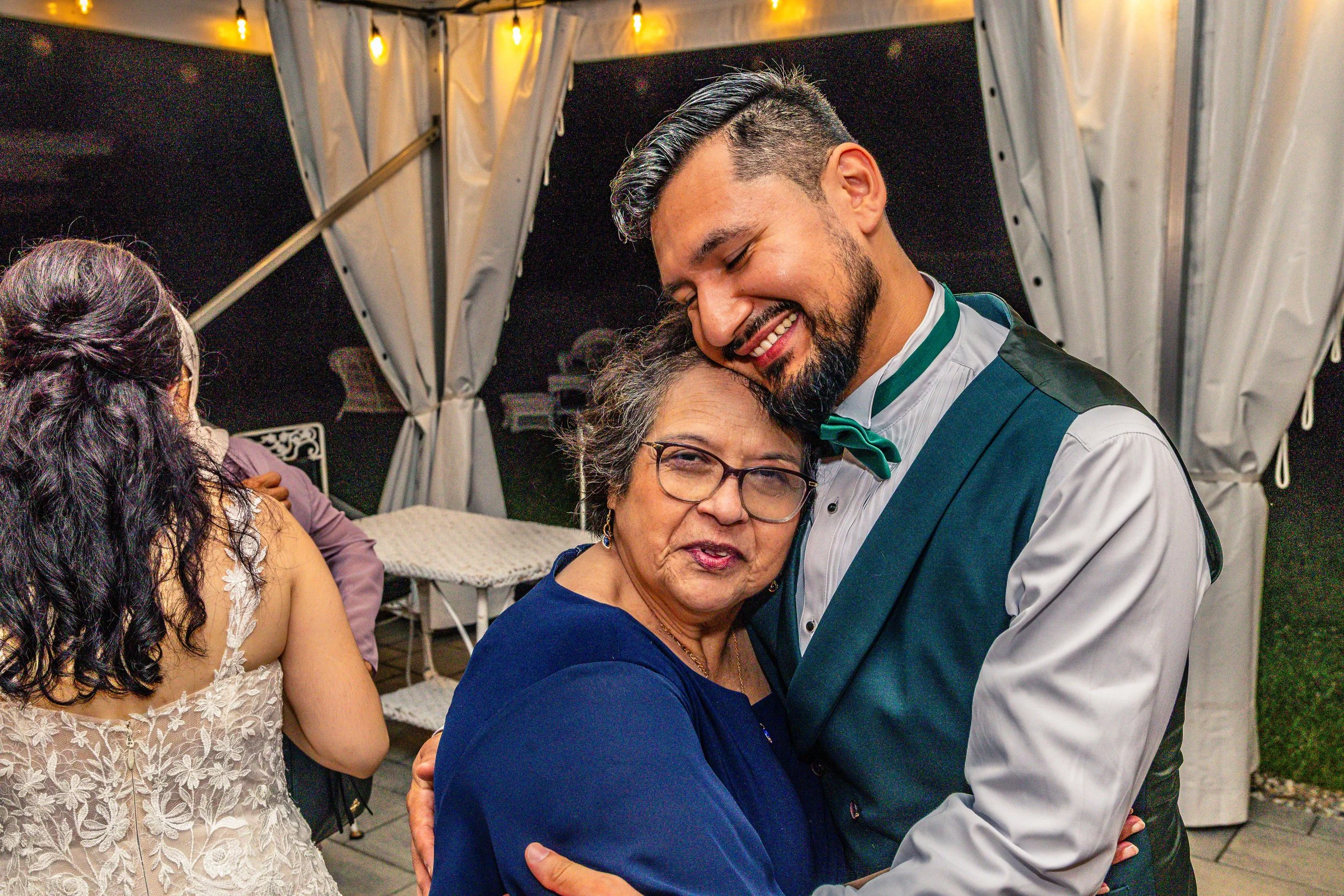 A man dressed in a tuxedo with a green bowtie and vest hugging an elderly woman with short gray hair and glasses at a gathering under a covered outdoor area at night.