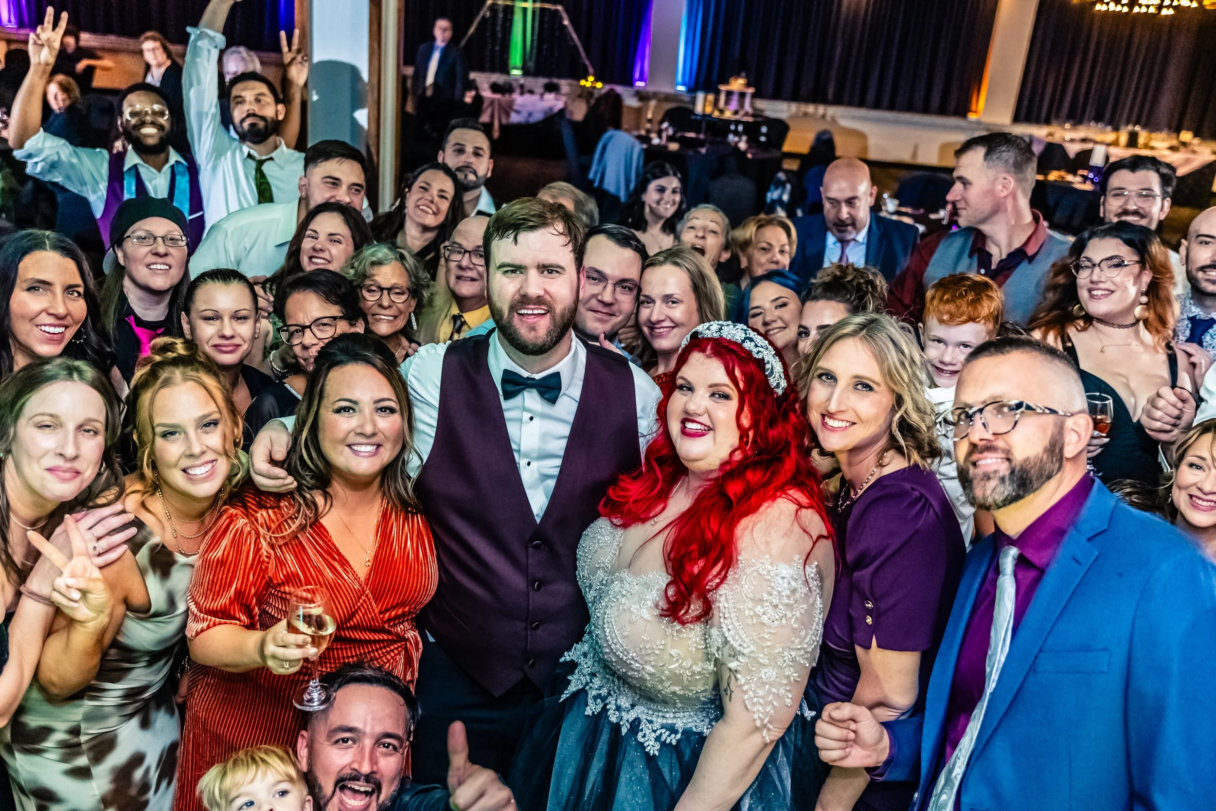 A large group of people at a wedding reception, including a bride with red hair and a groom in bow tie, smiling and posing for the camera in a festive indoor setting.