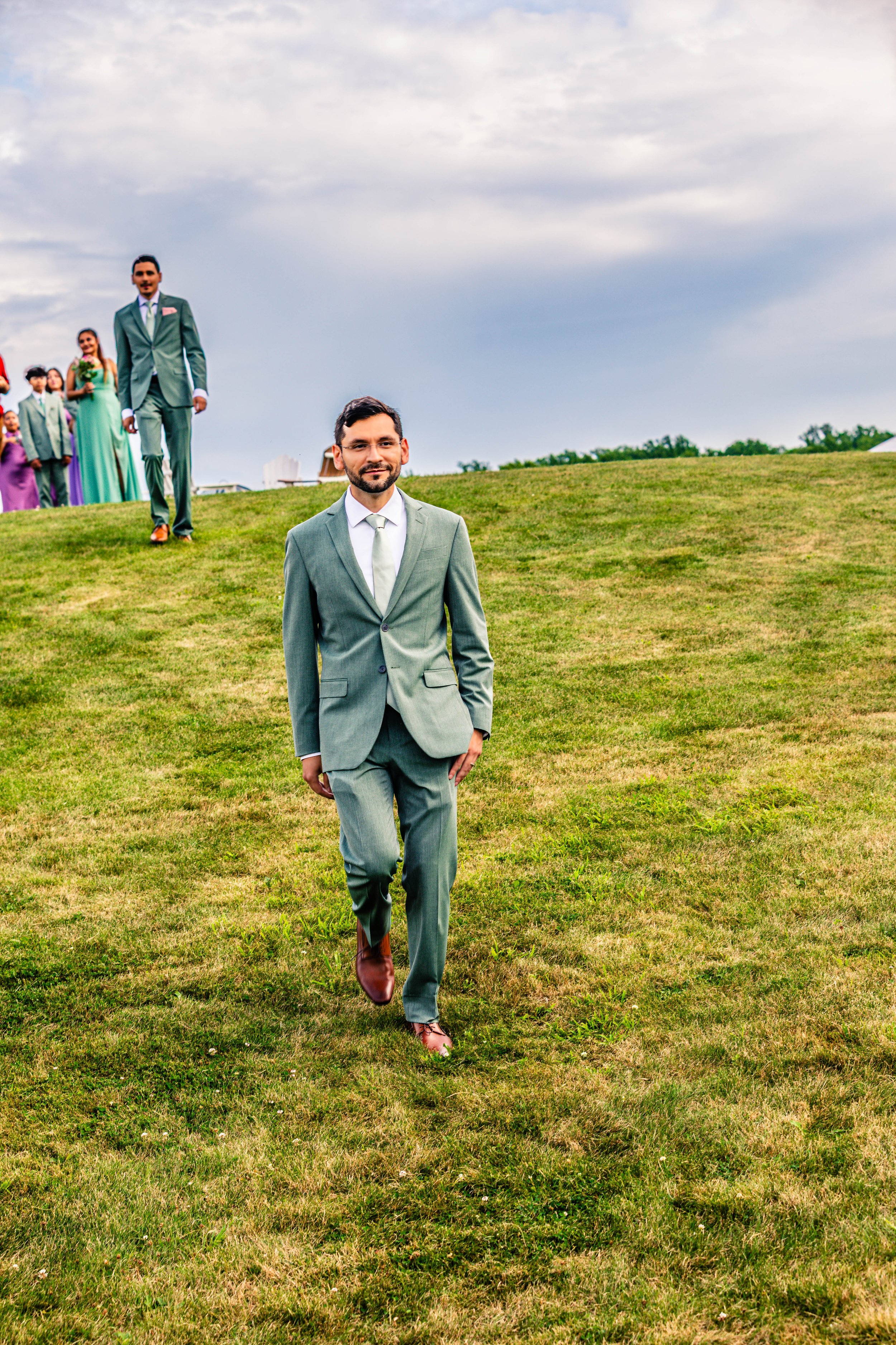 A man in a gray suit walking on a grassy hill with others dressed in colorful outfits in the background.