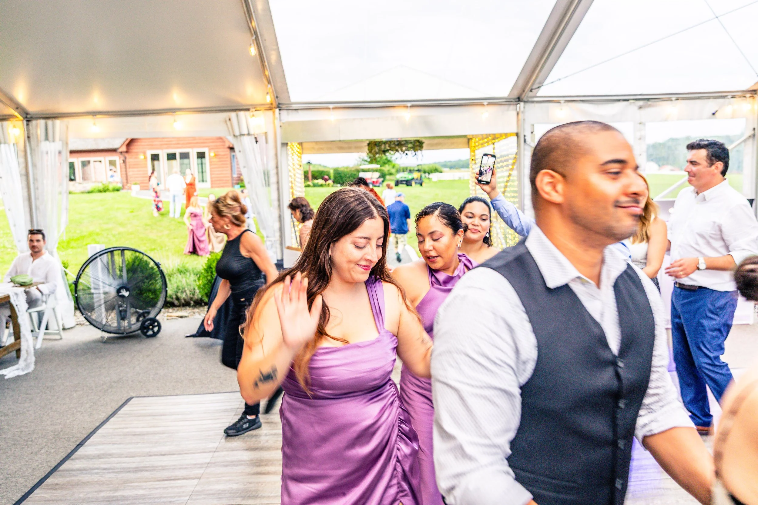 People dancing at a wedding reception inside a large outdoor tent, with a view of a grassy field and a barn in the background.