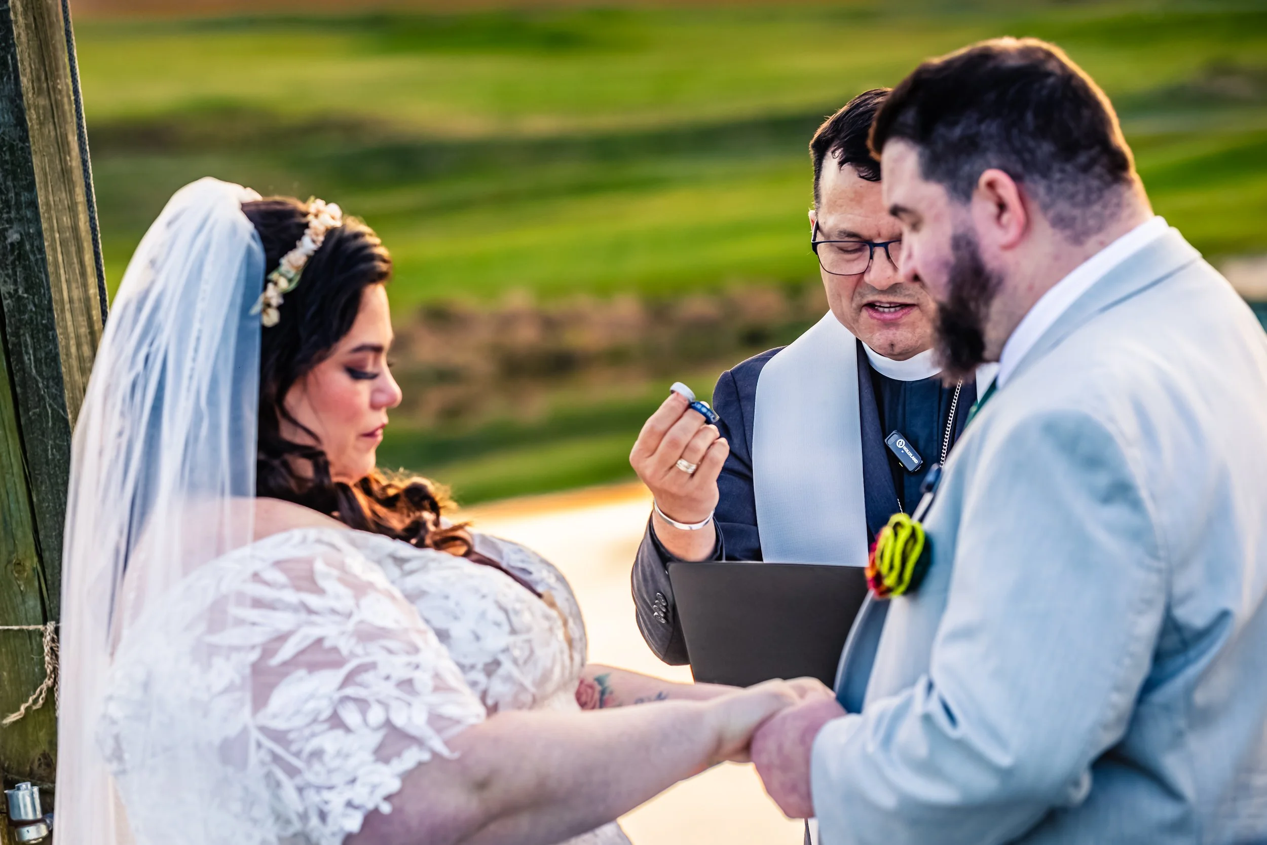 A bride and groom holding hands during their wedding ceremony outdoors, with an officiant reading a vows or prayer.