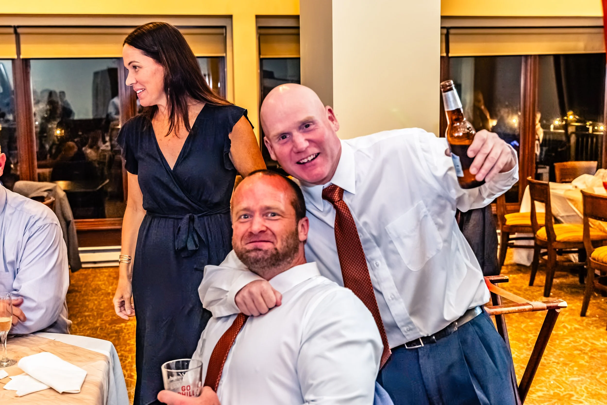 Three people at a dinner party, two men in white shirts and ties, one woman in a dark dress, with people and windows in the background.