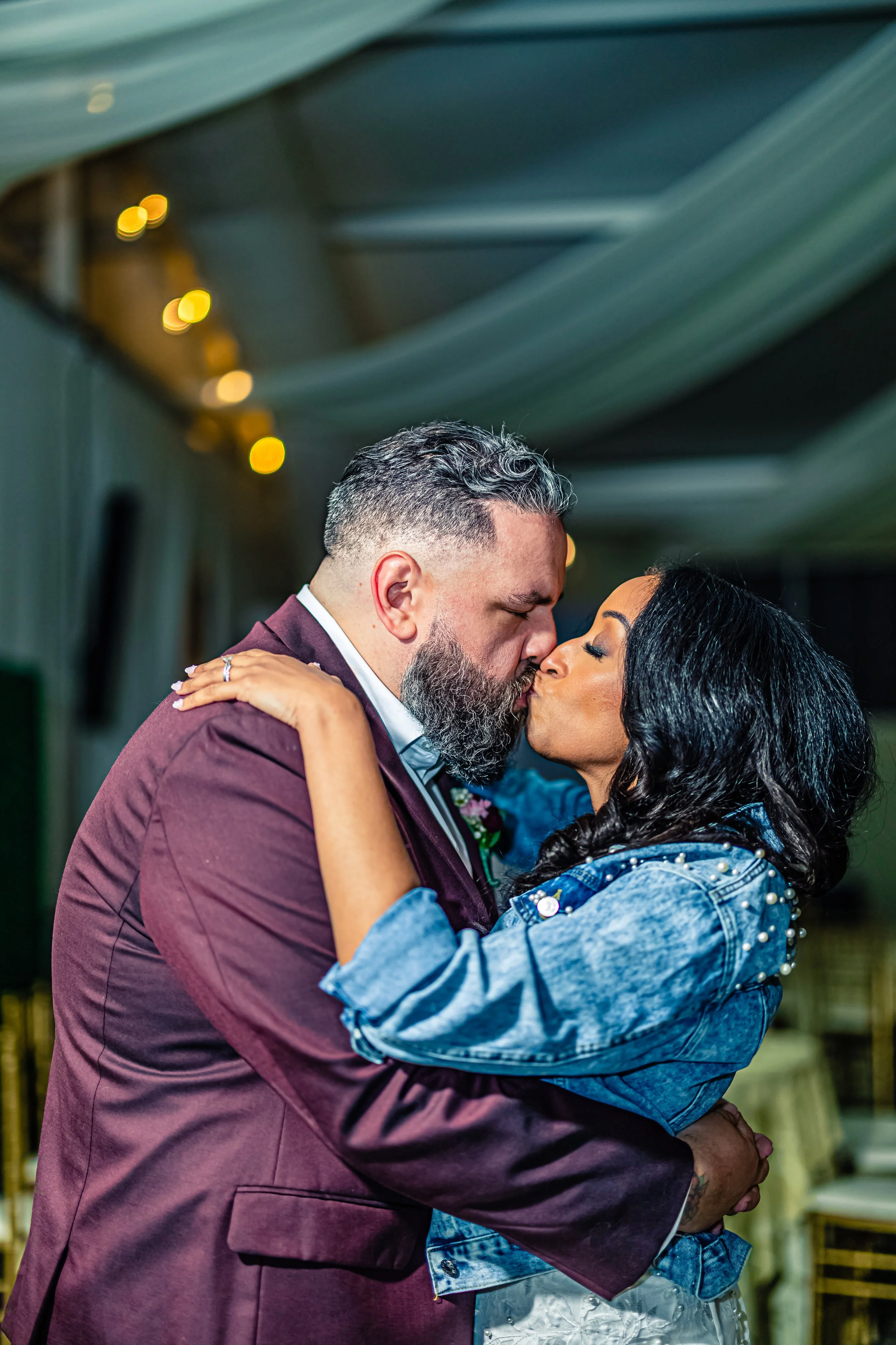 A man with a beard and gray hair and a woman with black hair are kissing at a wedding reception.