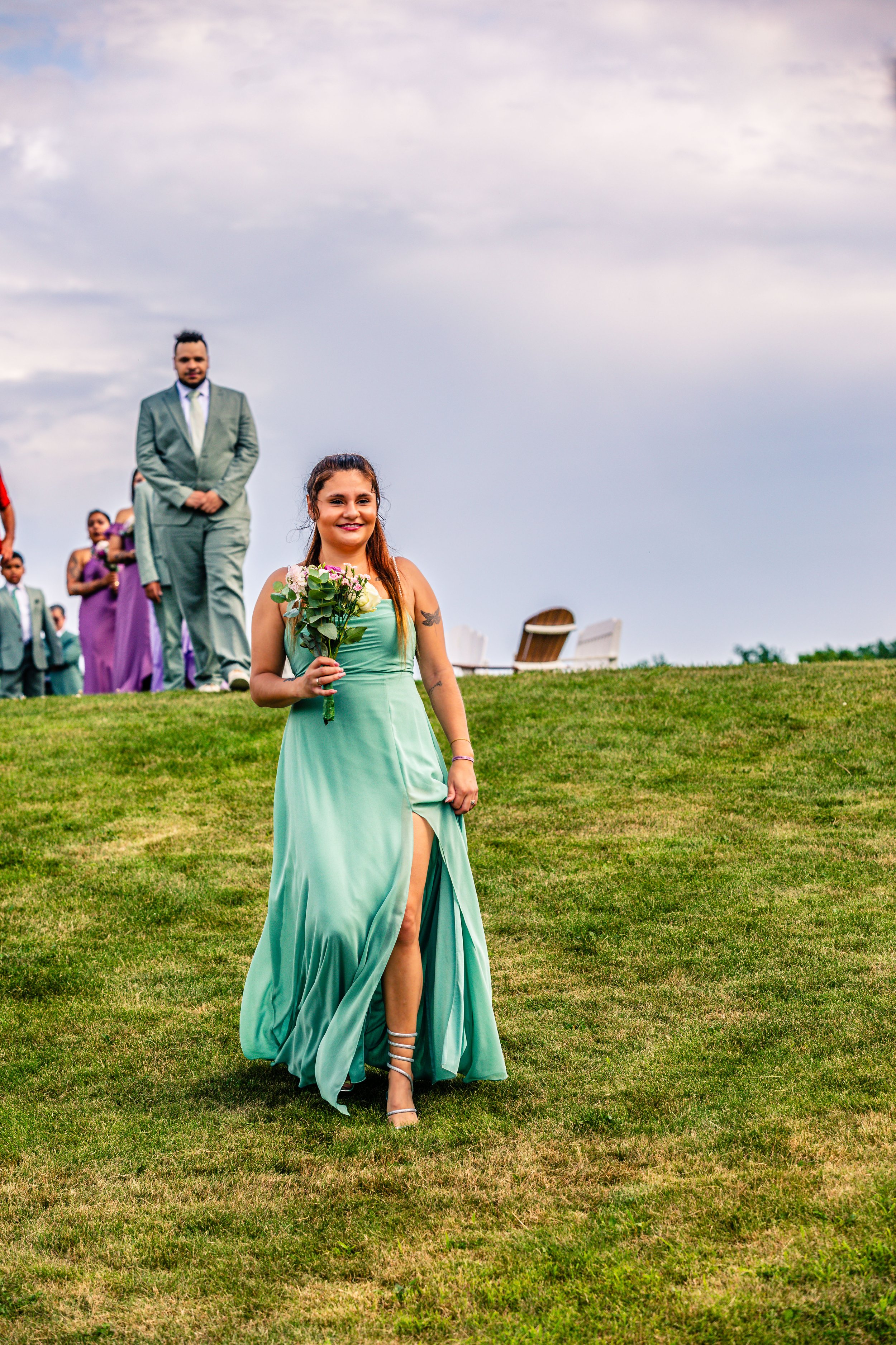 A young woman in a green dress holding a bouquet of flowers, smiling, with a group of people in formal attire walking behind her on a grassy hill during daytime.