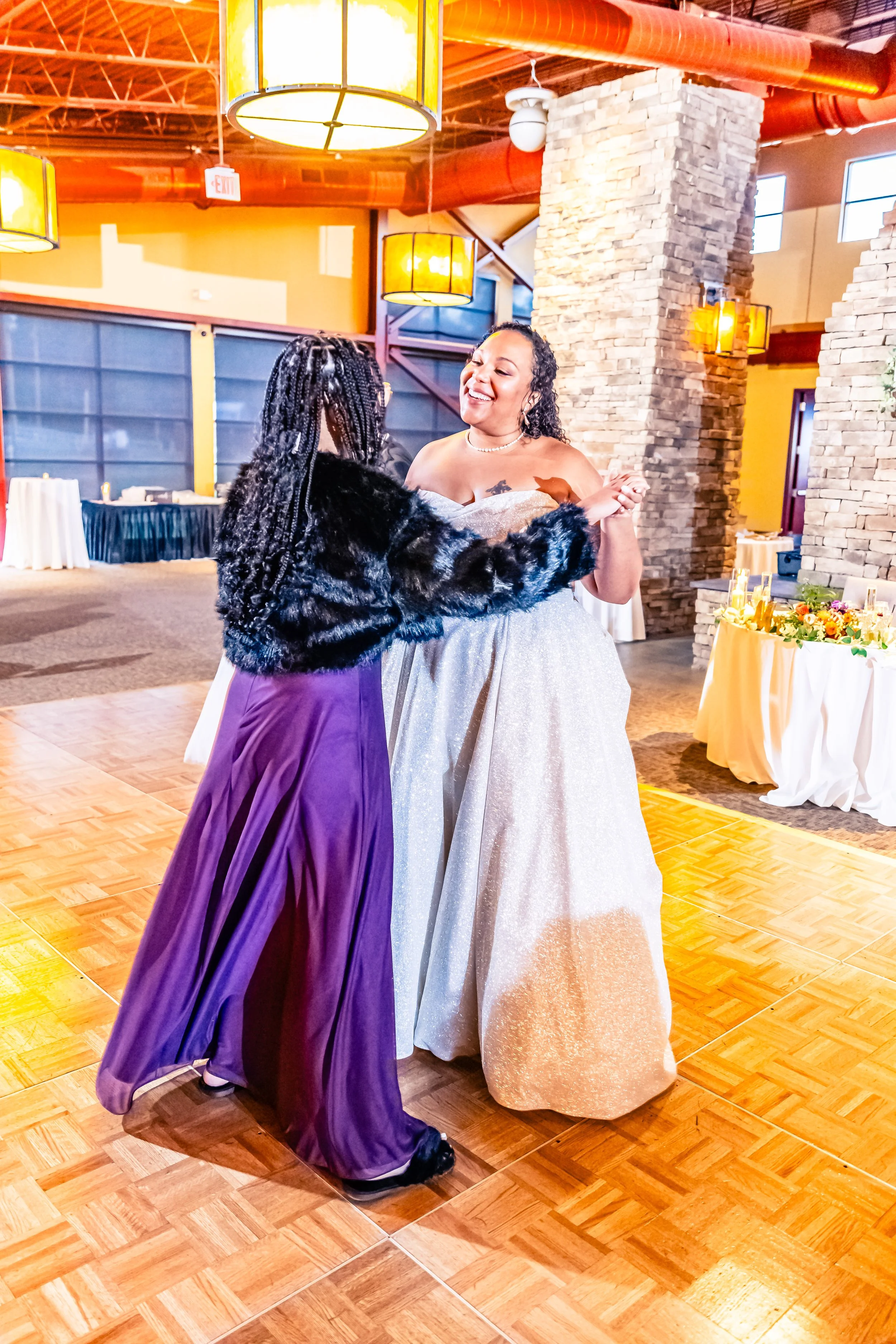 Two women dancing at a wedding reception, one in a sparkly dress and the other in a purple dress with a black faux fur jacket, inside a decorated banquet hall.