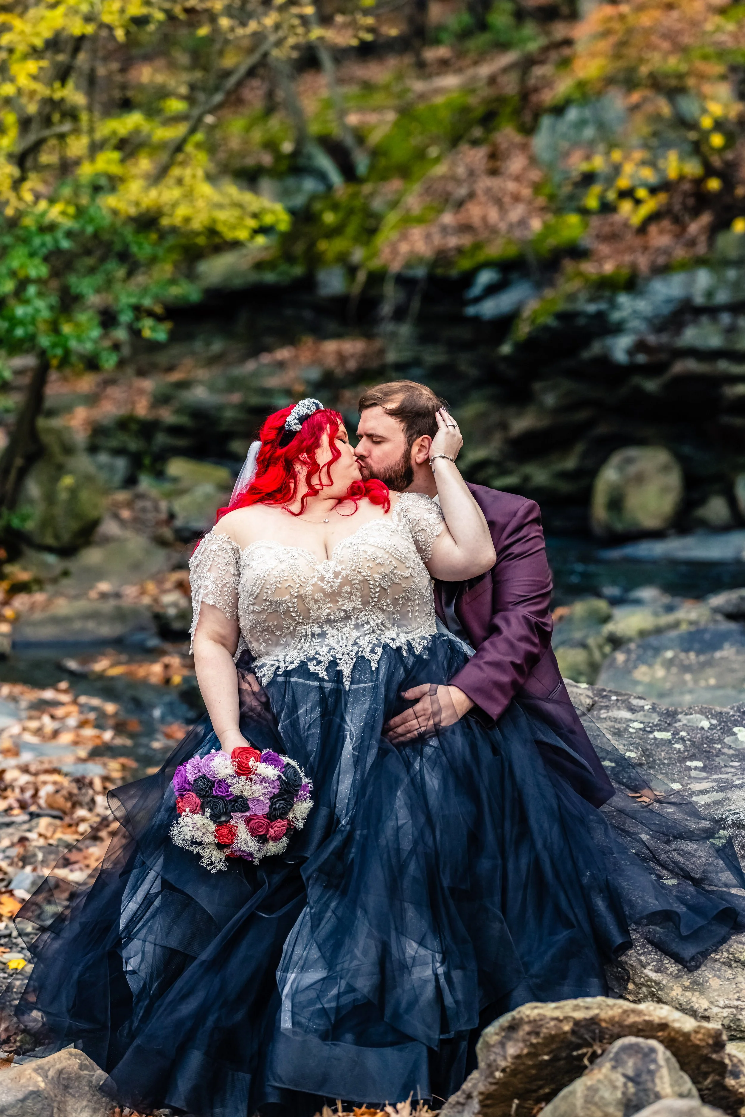 Couple in wedding attire sharing a kiss outdoors near a creek, with trees and rocks in the background.