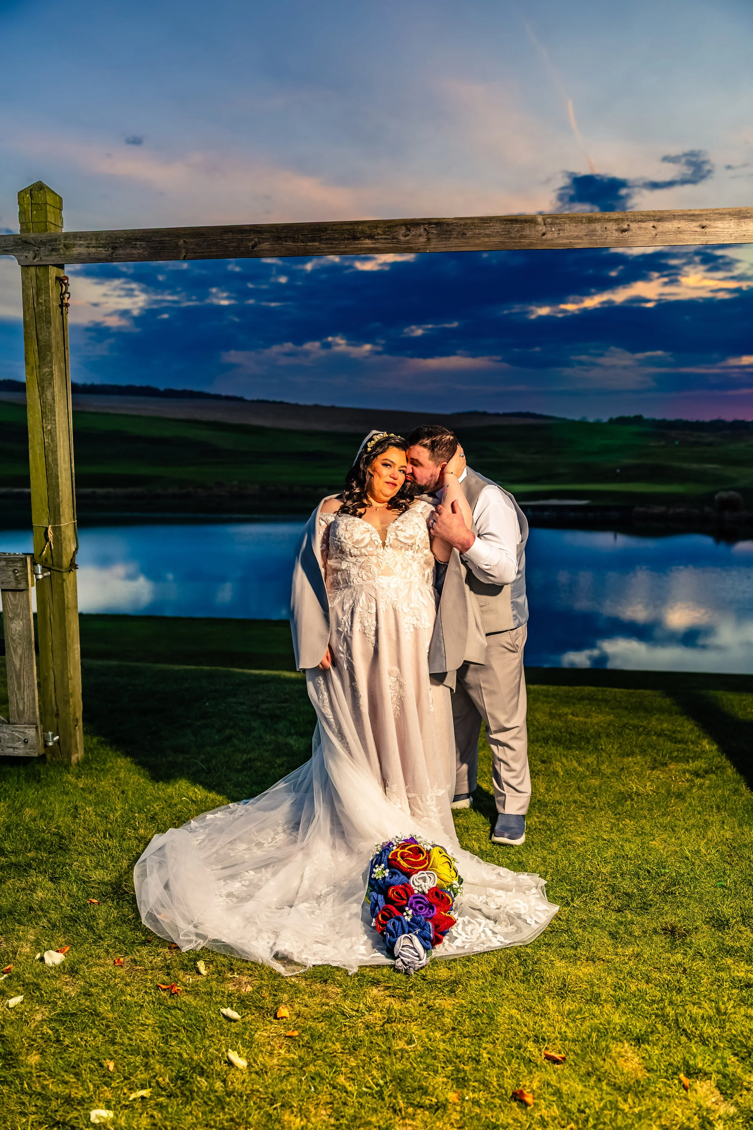 A newlywed couple embracing outdoors at sunset, with a lake and sky in the background, and a colorful bouquet on the grass.