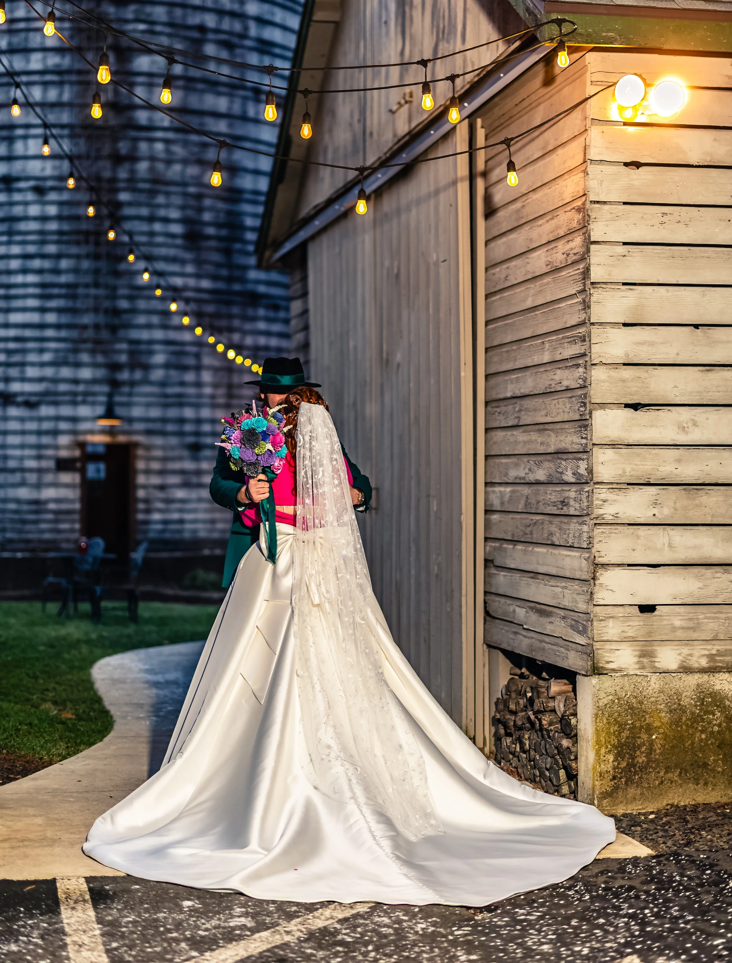 A bride and groom embrace outside a rustic building under string lights at dusk. The bride is wearing a long white wedding gown with a veil, and the groom is dressed in dark clothing with a hat. The groom holds a bouquet of colorful flowers.