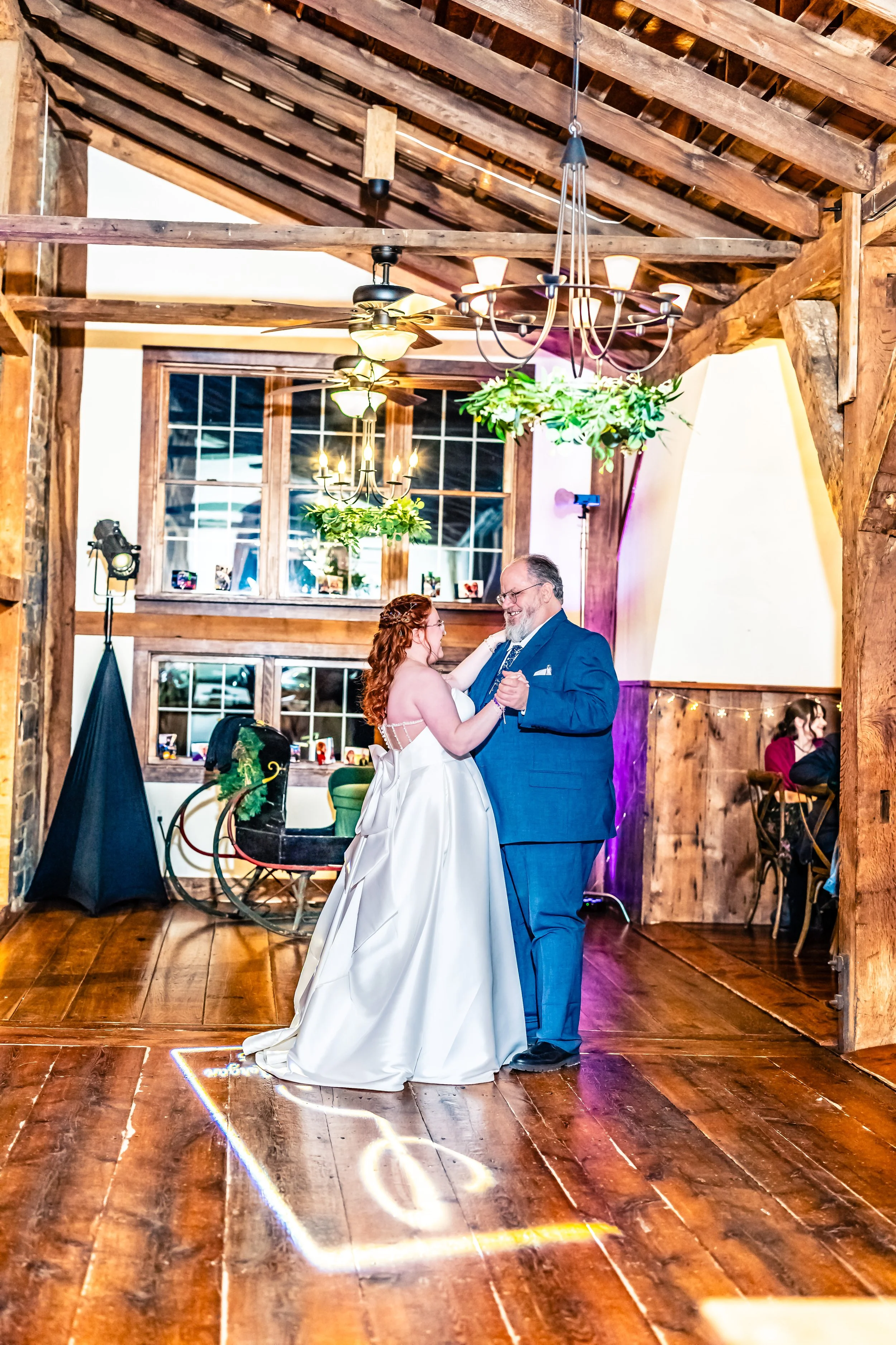 A bride and groom dancing in a rustic reception hall with wooden beams, chandeliers, and large windows.