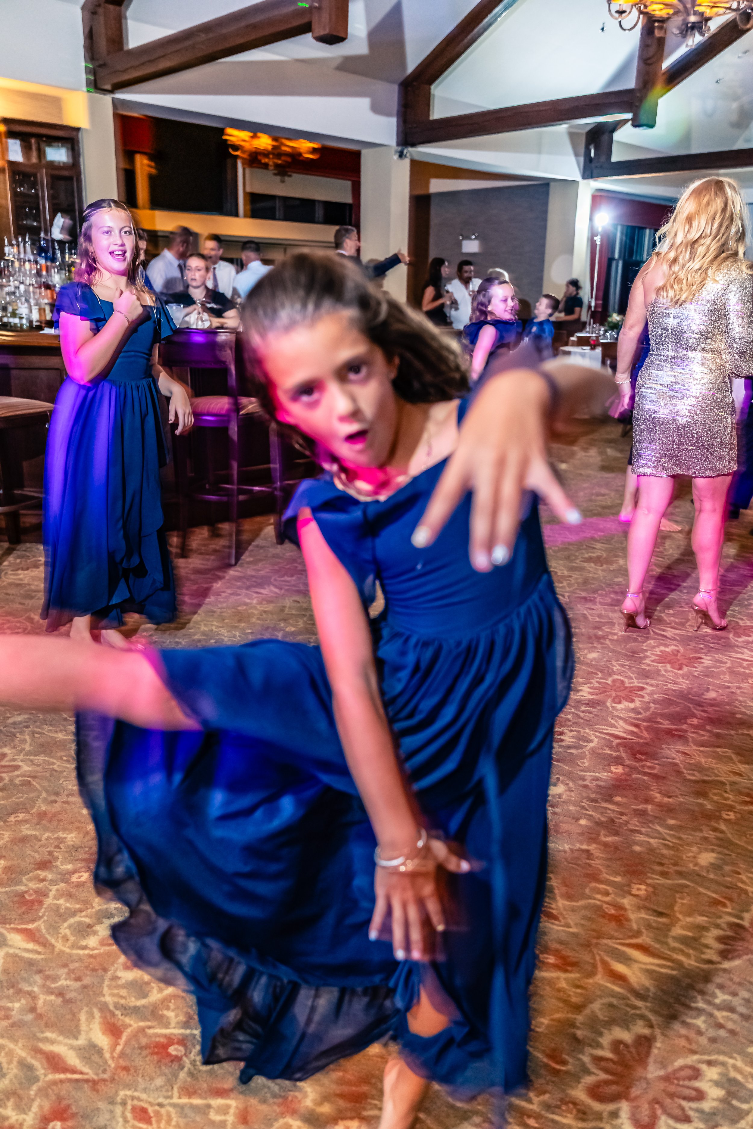 A young girl in a blue dress dancing and striking a pose at a lively party or dance event, with other children and adults in the background.