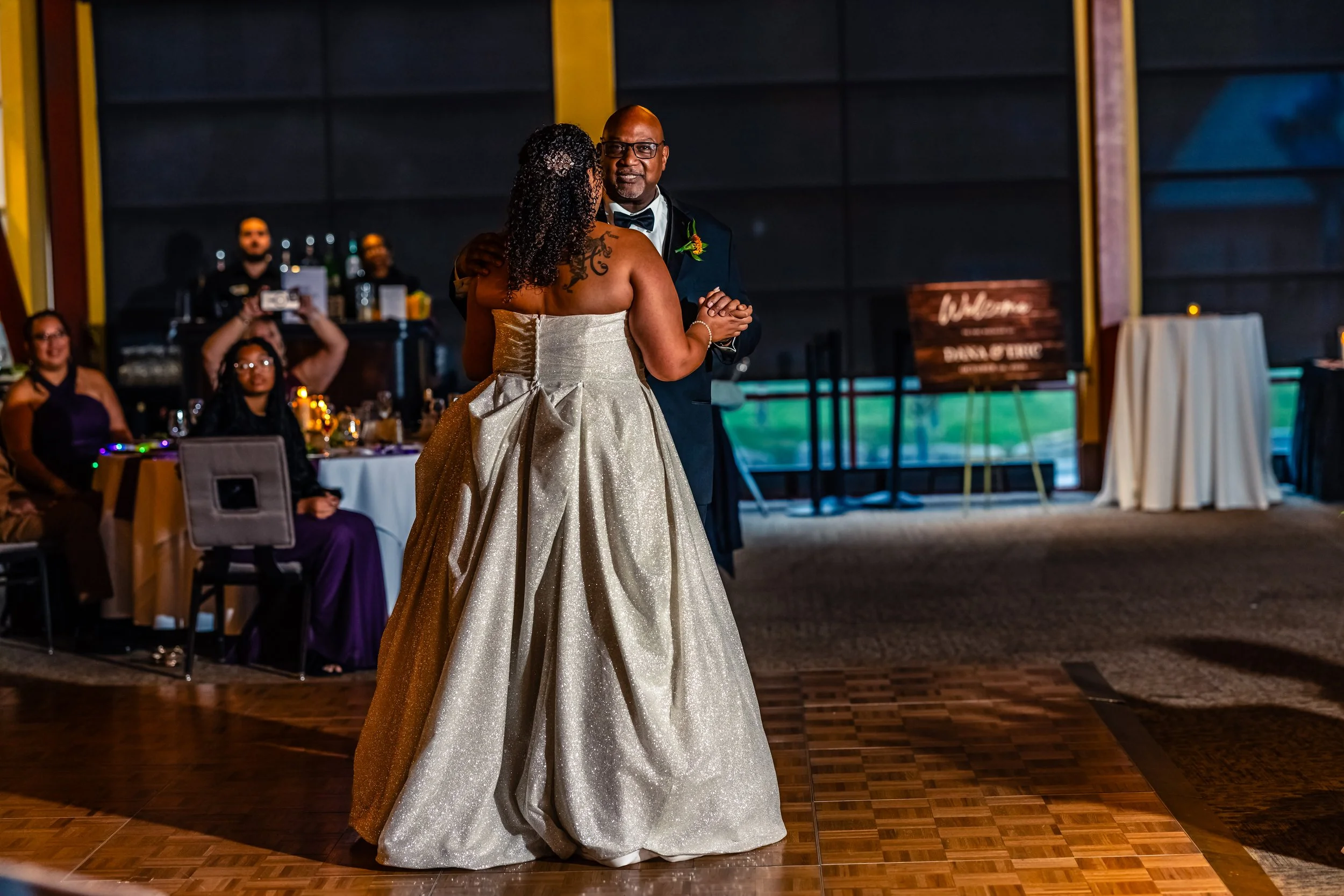A bride and groom dancing at their wedding reception, with guests watching and smiling in the background.