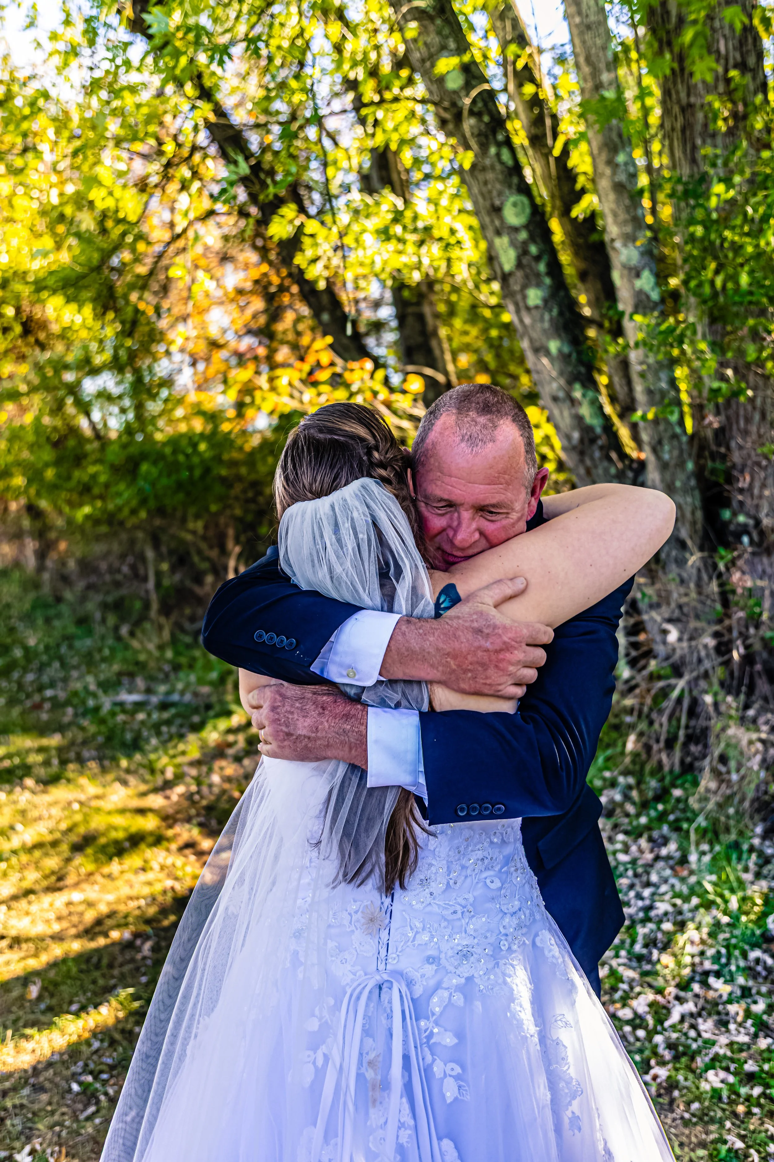 A bride and a man, possibly her father, sharing an emotional hug outdoors with trees and sunlight in the background.