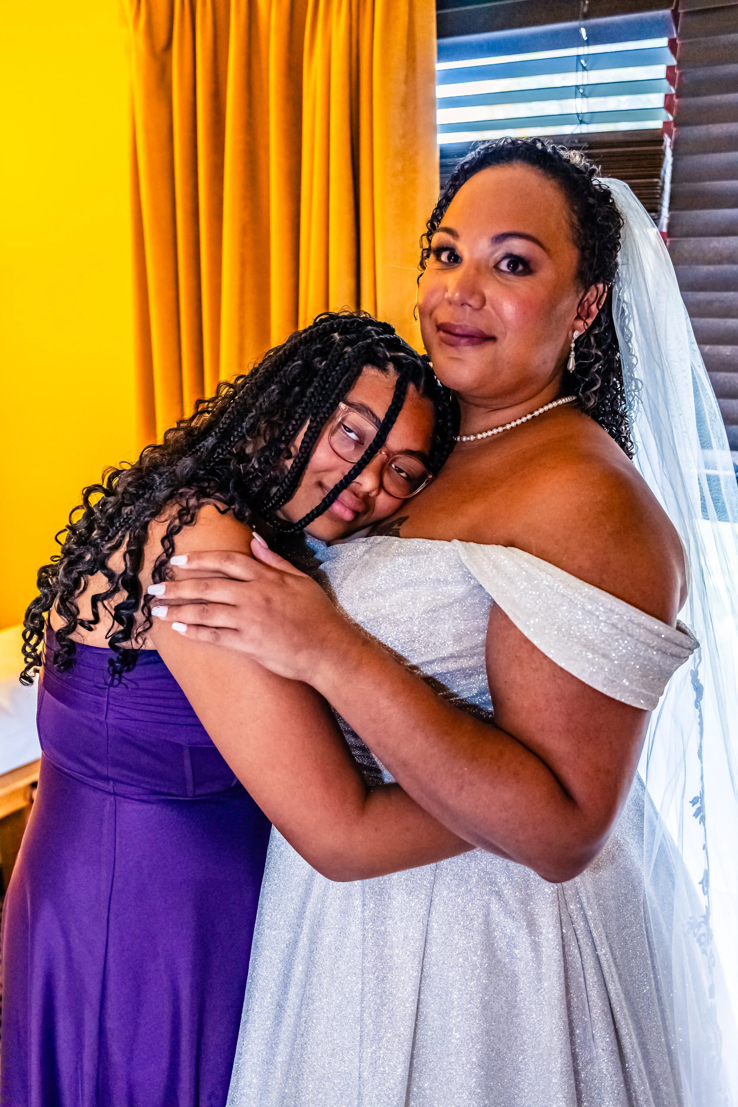 A bride in a wedding dress hugging a woman in a purple dress, both smiling and looking at the camera in a room with yellow curtains and a window with blinds.