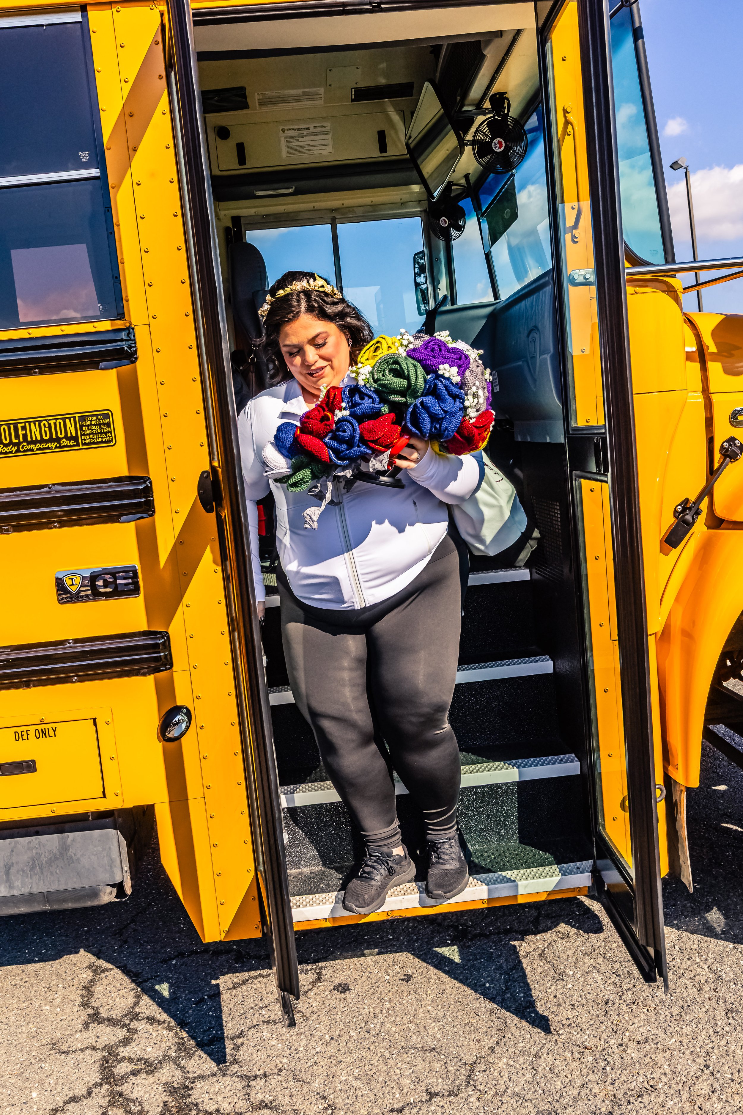 A woman with dark, curly hair stepping off a yellow school bus holding a colorful bouquet of knitted flowers, wearing a white jacket and black leggings.