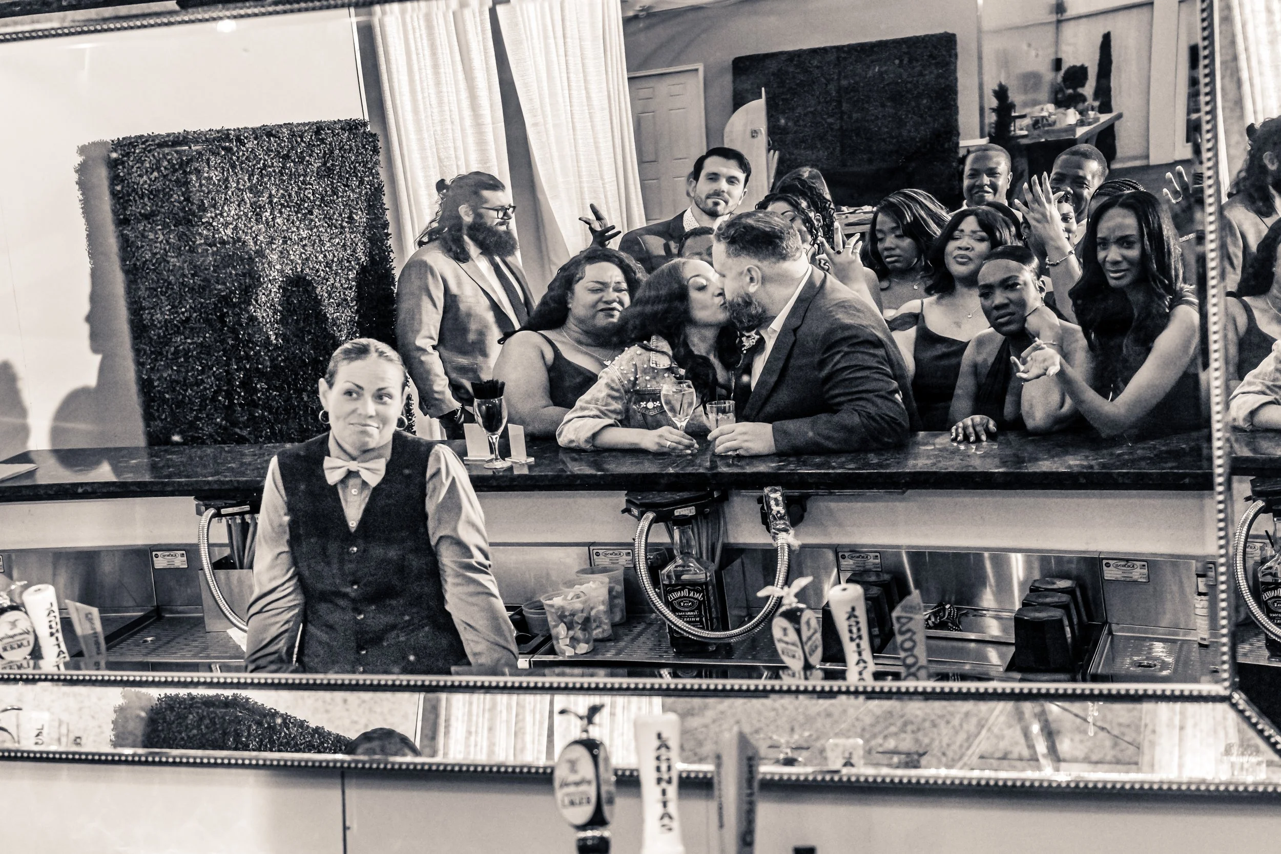 A black-and-white photo of a crowded bar with a bartender woman in the foreground and a photo of a big gathering of people in the background.