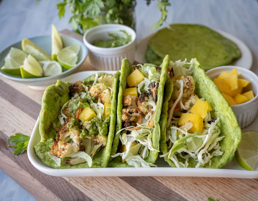 A rectangular plate holds three tacos made from spinach tortillas stuffed with roast avocado, mango, shredded cabbage, and chimichurri sauce.