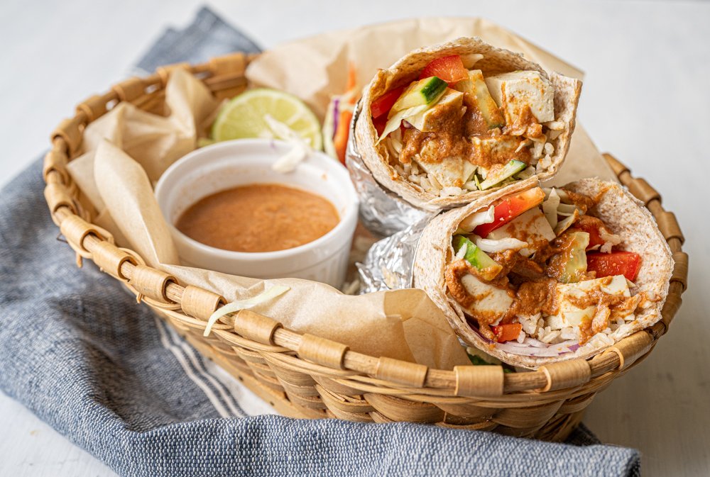 Two tofu and vegetable wraps are in a wooden basket, one on top of the other, along with a small ramekin of chipotle dressing and a half lime.