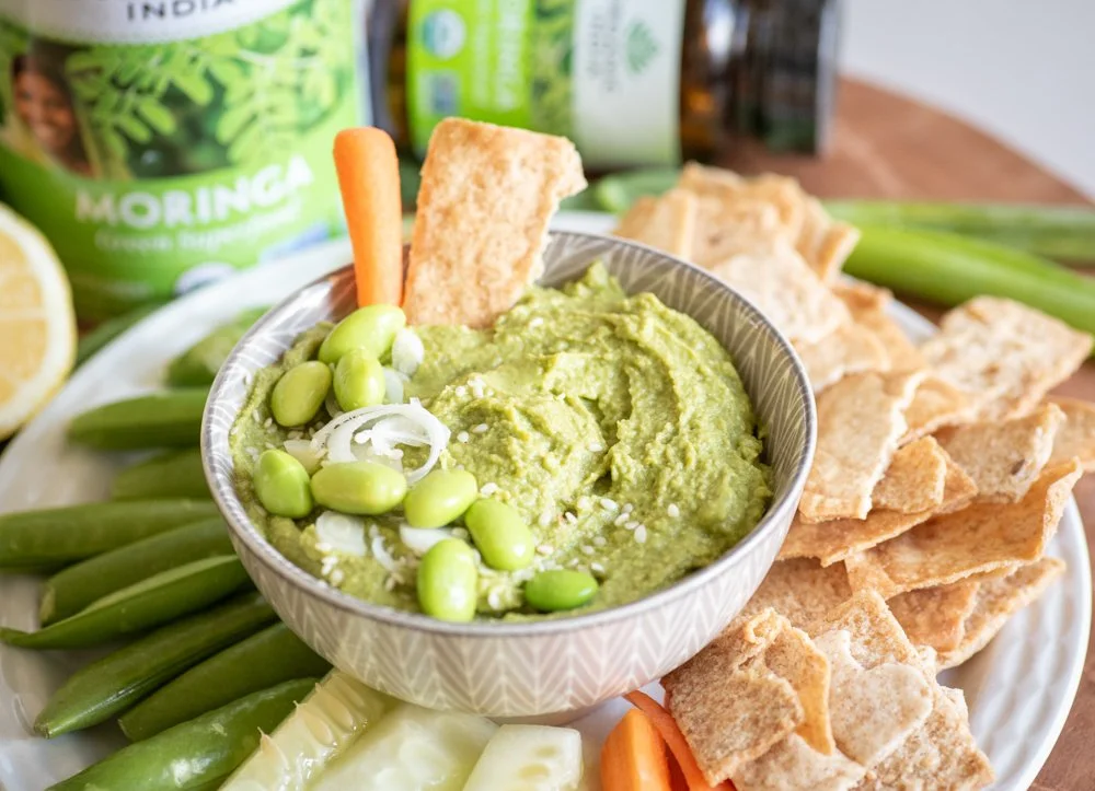 A small bowl of edamame dip viewed as if seated at a table; garnished wiuth edamame, green onions, and sesame seeds and centered on a plate surrounded byu pita chips, carrot and cucumber sticks, and snap peas.