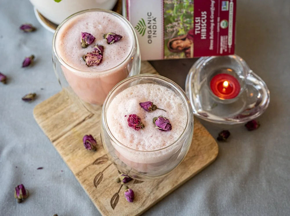 An overhead view of two frothy pink hibiscus tea lattes. Dried mini roses are sprinkled around, and a glass heart-shaped votive with a red candle is lit alongside.