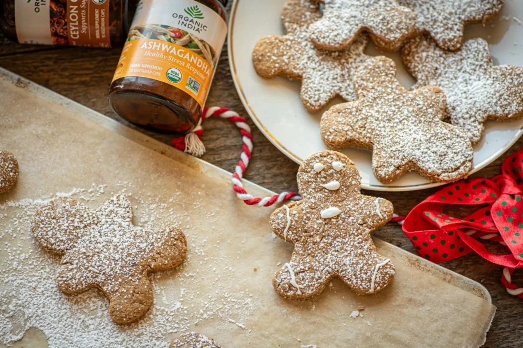 Some gingerbread cookies dusted with powdered sugar are on a place next to a baking sheet with cookies on parchment paper. One gingerbead man cookie is halfway on the plate, smiling.