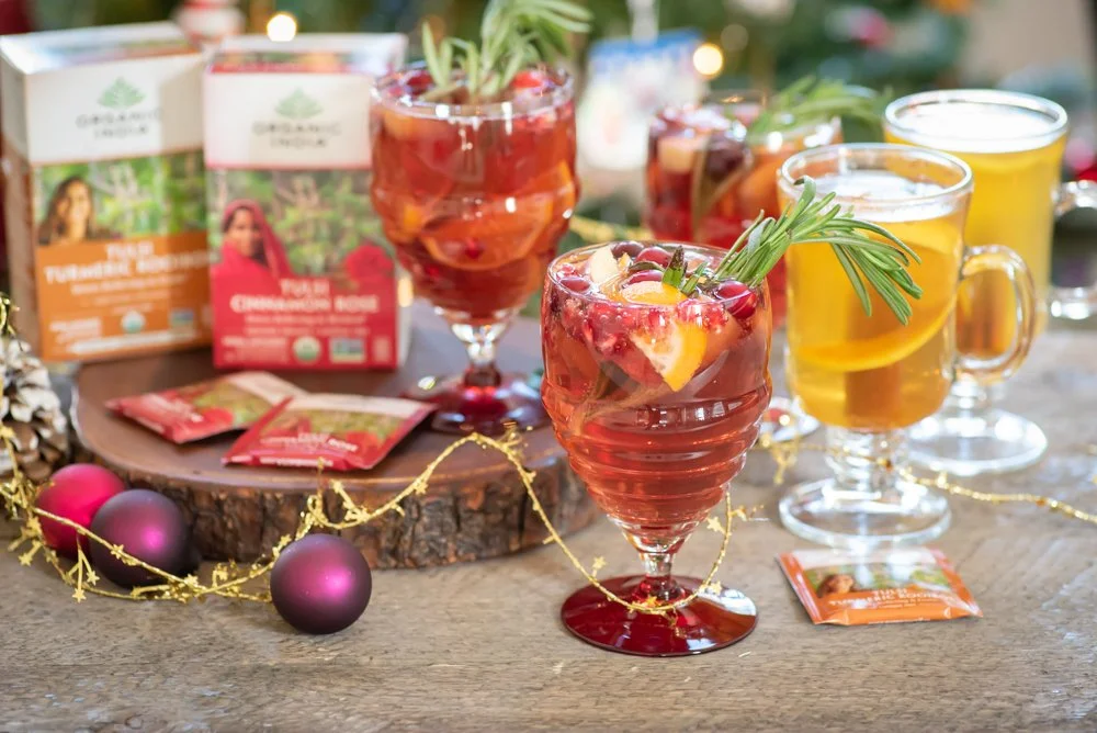 Assorted mocktails are placed on a wooden table with a background of holiday lights, tea packets and boxes, and decorations.