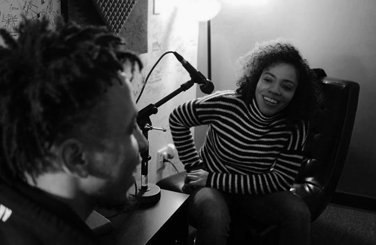 A woman with curly hair smiling and talking to a man with locks in a recording studio with microphones.