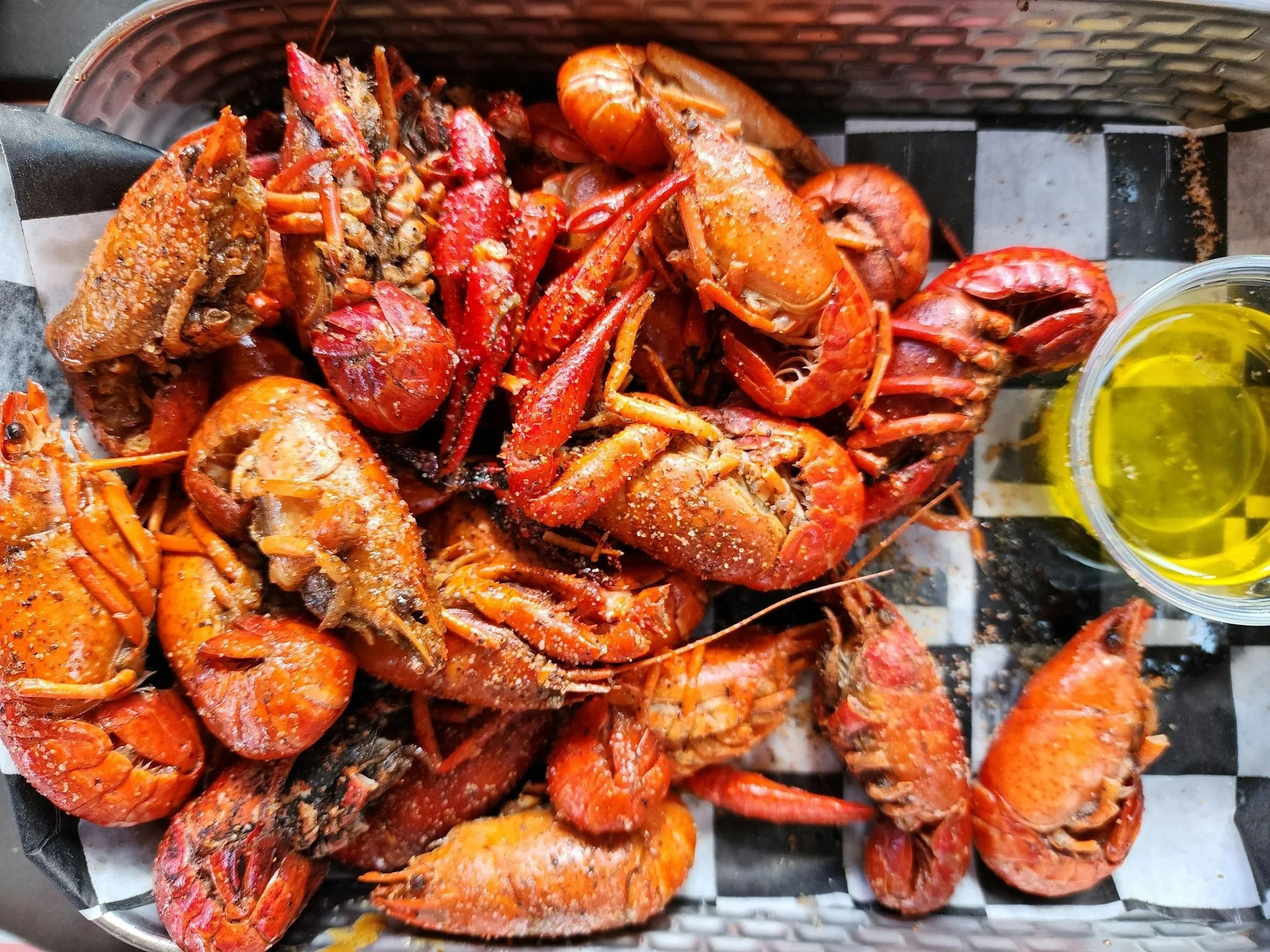 A collection of cooked lobsters with salt on their shells, served on a checkered black and white tray with a small container of melted butter.