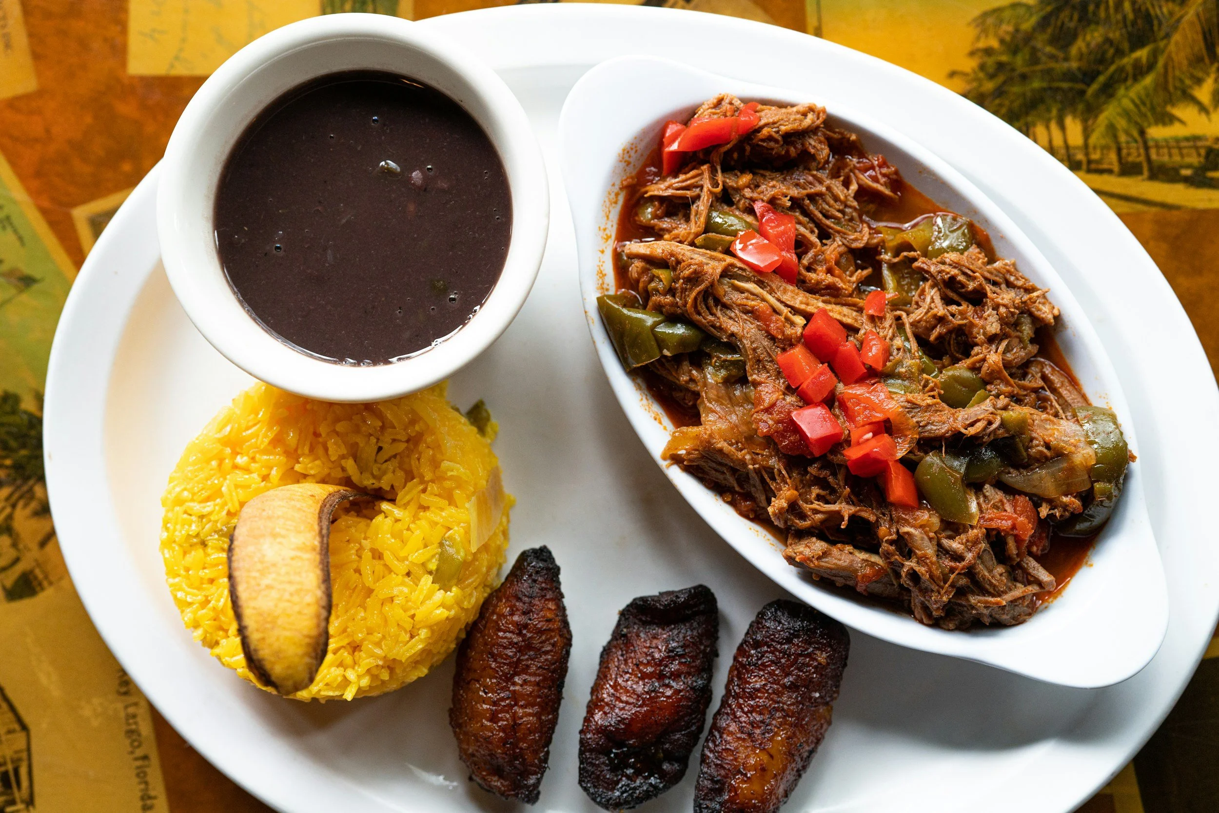 Plate of Latin American food including yellow rice, shredded beef stew with vegetables, grilled plantains, and fried meatballs, with a cup of black coffee or thick sauce.
