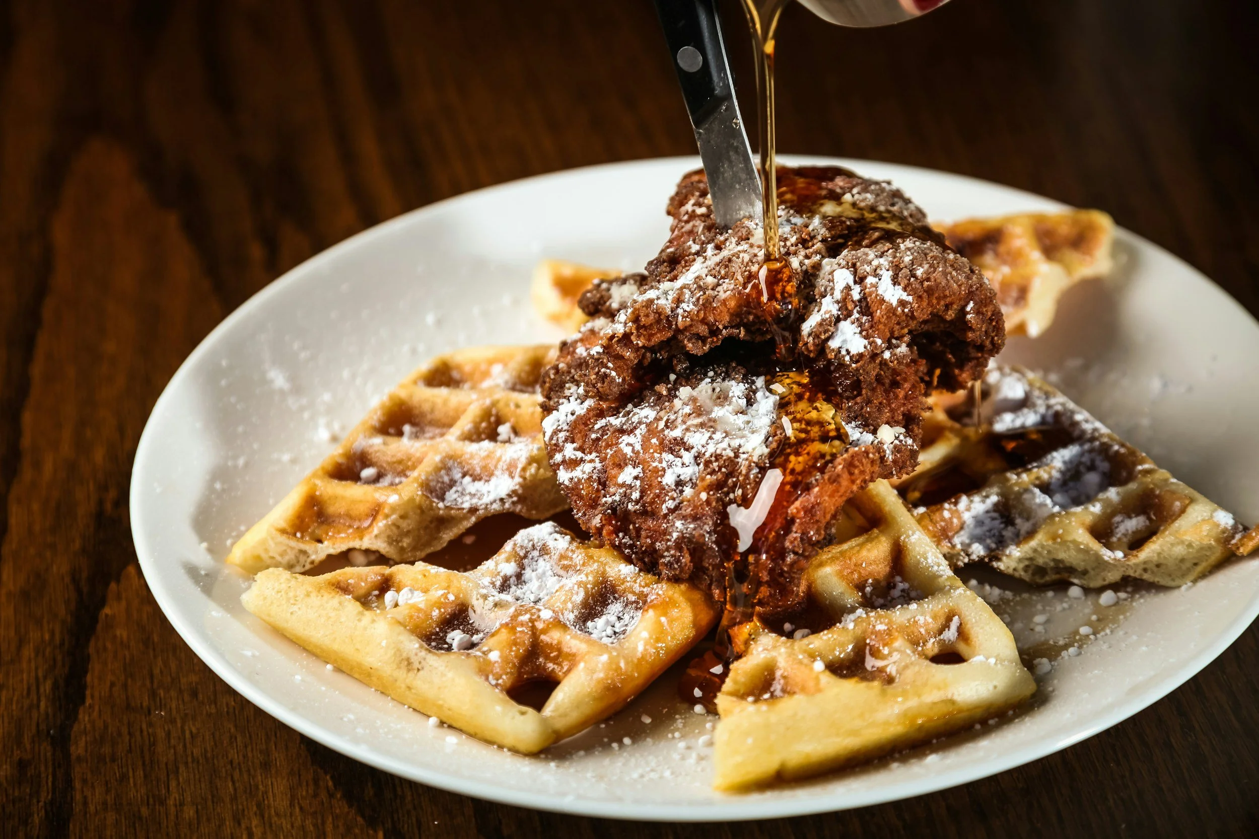 Close-up of a waffle topped with a fried chicken drumstick, drizzled with syrup, and dusted with powdered sugar, served on a white plate.