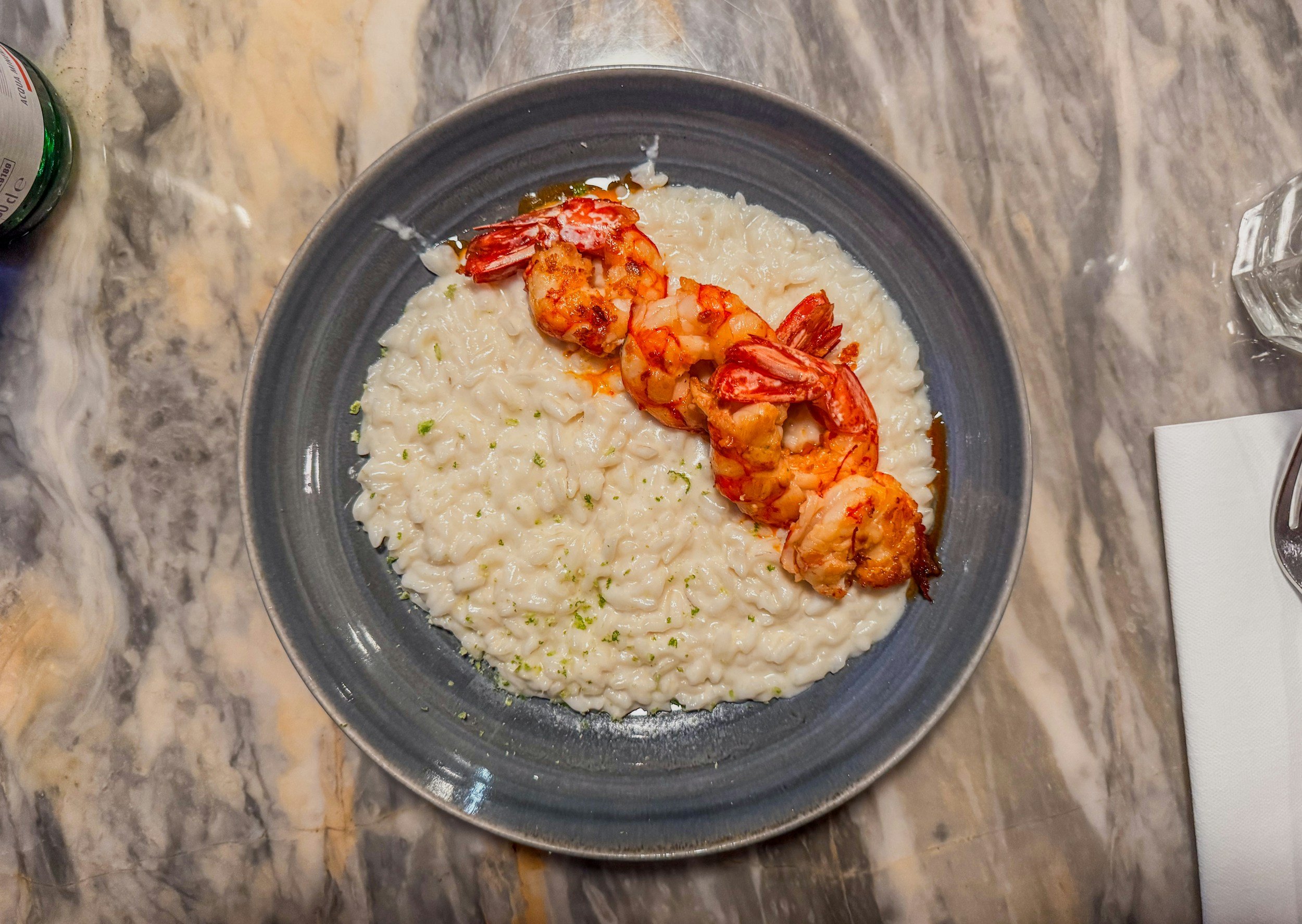 A plate of creamy risotto garnished with lime zest and topped with grilled shrimp on a dark ceramic dish, on a marble countertop.