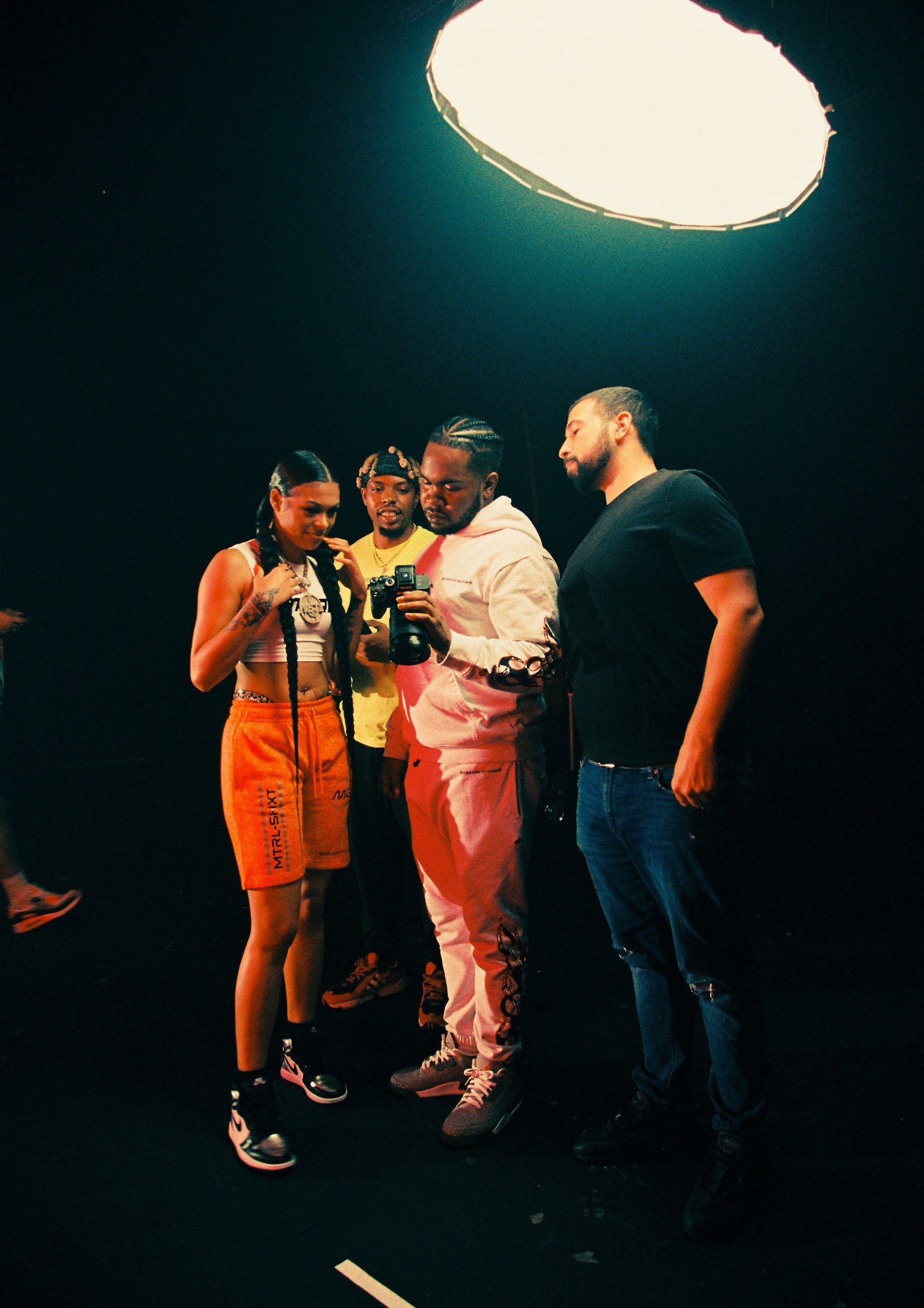 Four young adults standing together under a large overhead light in a dark studio, looking at a camera or phone held by one of them, with black background and casual attire.