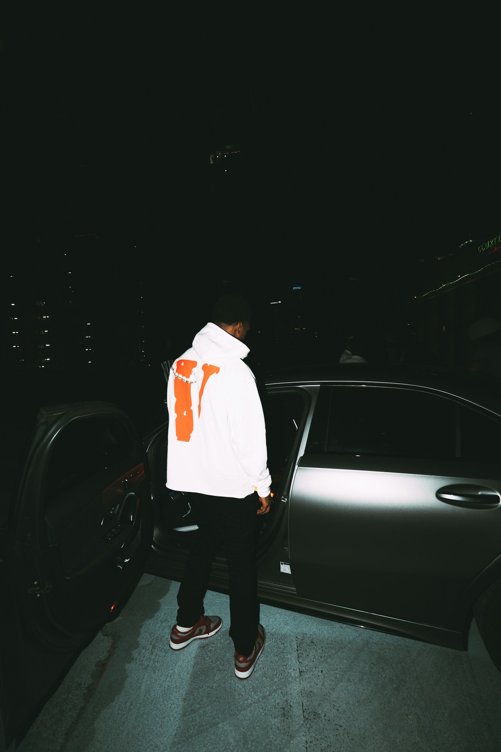 A man wearing a white hoodie with orange design, black pants, and red and white sneakers is standing next to a dark-colored car at night. He appears to be opening or closing the car door.