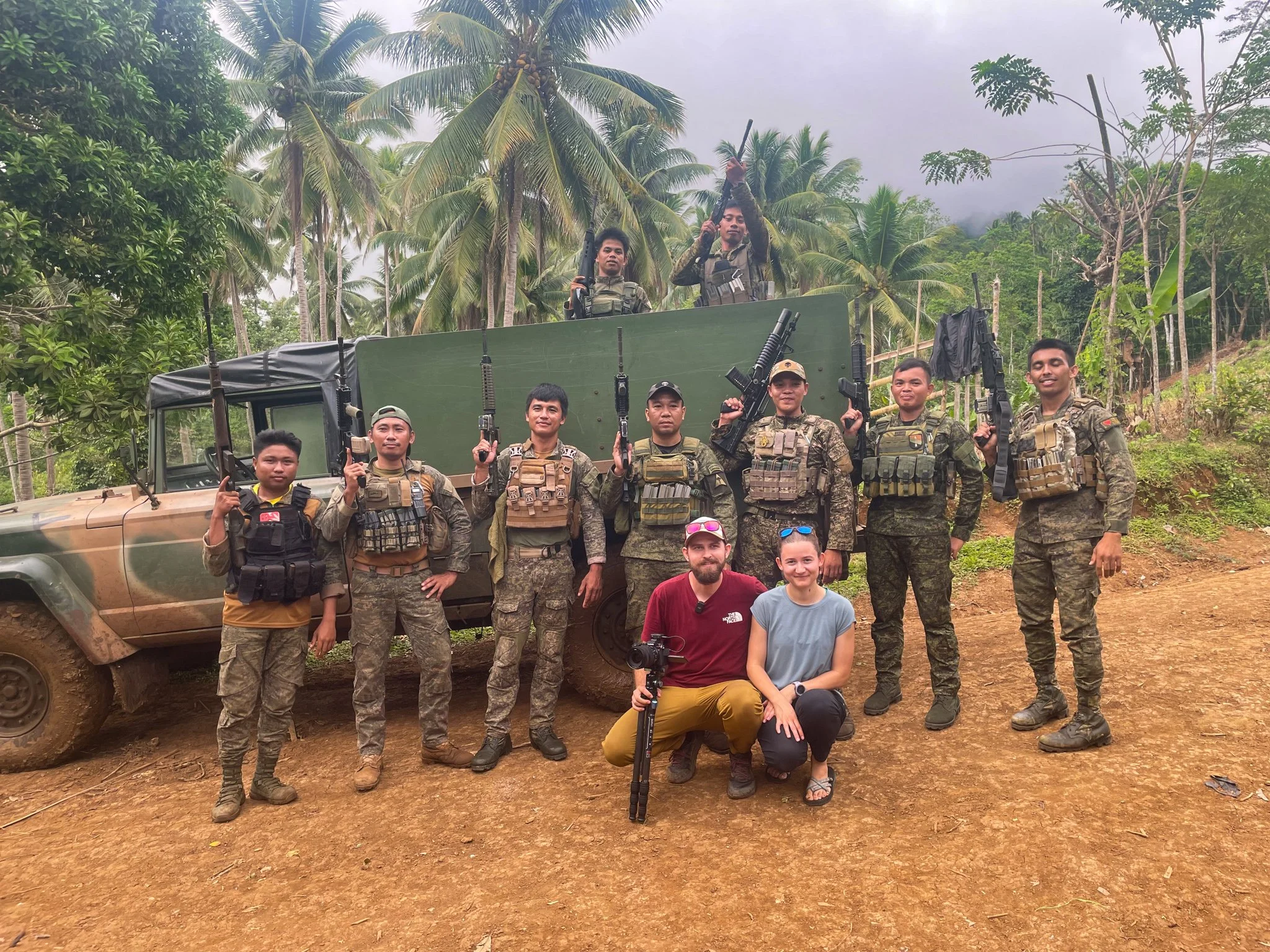 Group of soldiers in camouflage uniforms holding guns, standing in front of a military vehicle on a dirt path with palm trees and jungle surroundings, two civilians seated in front, with two soldiers on top of the vehicle