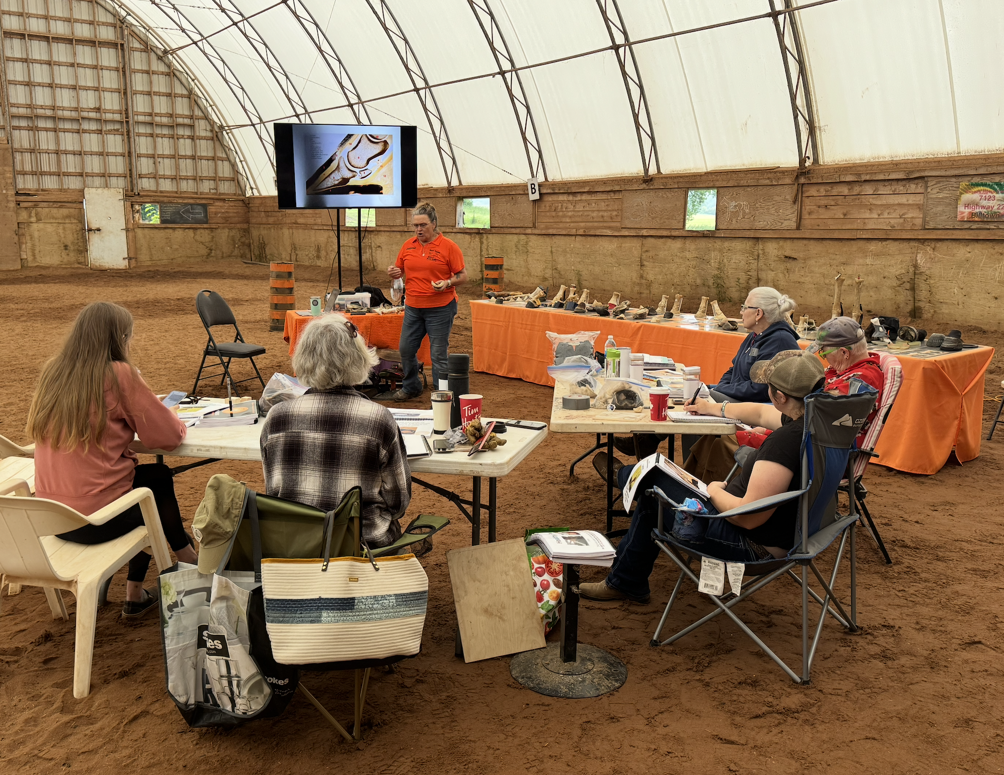 A woman is giving a presentation in an indoor riding arena with a dirt floor. She stands in front of a large screen, displaying an image, while seated attendees take notes. There are tables with various items, including shoes and notebooks, and some chairs are stackable or foldable.