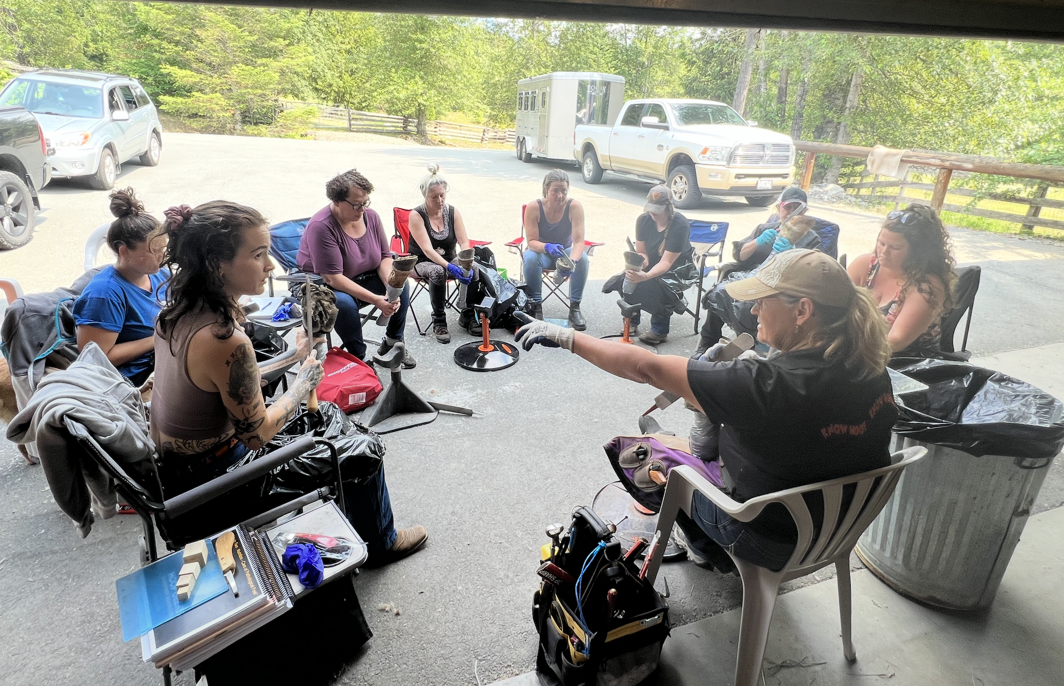 Group of women participating in a hoof geeks hoof care clinic outdoors, seated in a semi-circle with tools and supplies on their laps and tables, under a shaded area with trees and parked vehicles in the background.