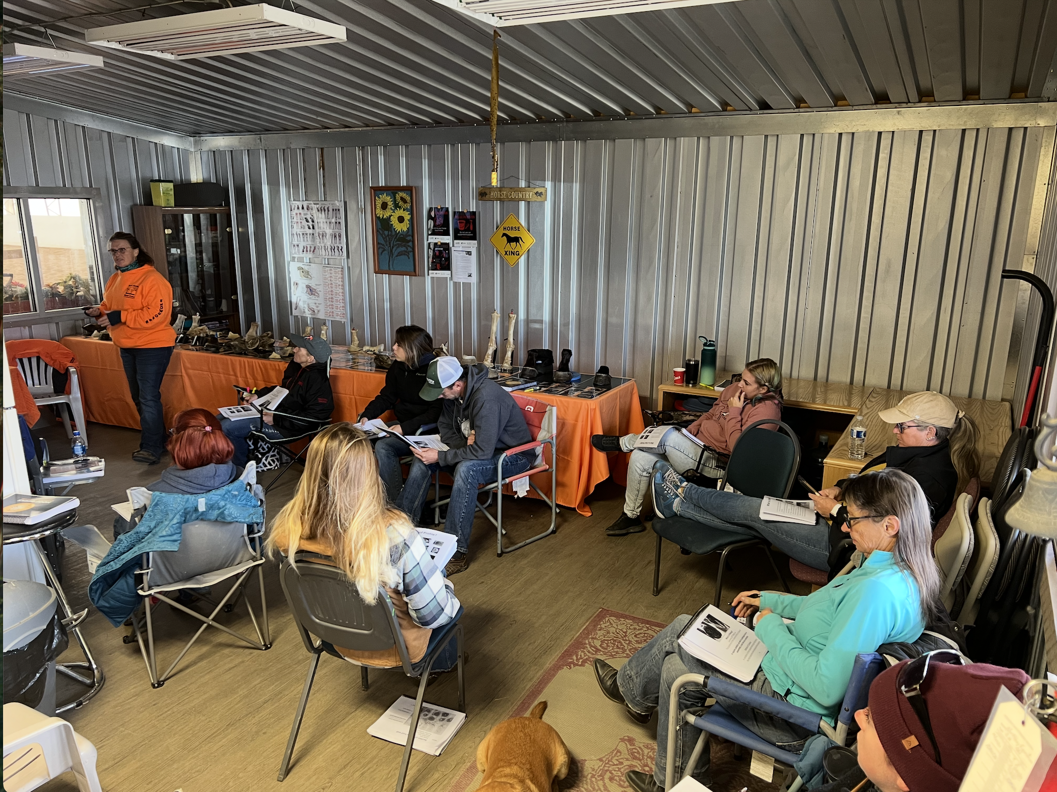 People seated in a room attending a presentation or meeting, some reading or listening while a woman in an orange sweatshirt addresses the group, with animal bones and posters on the wall.