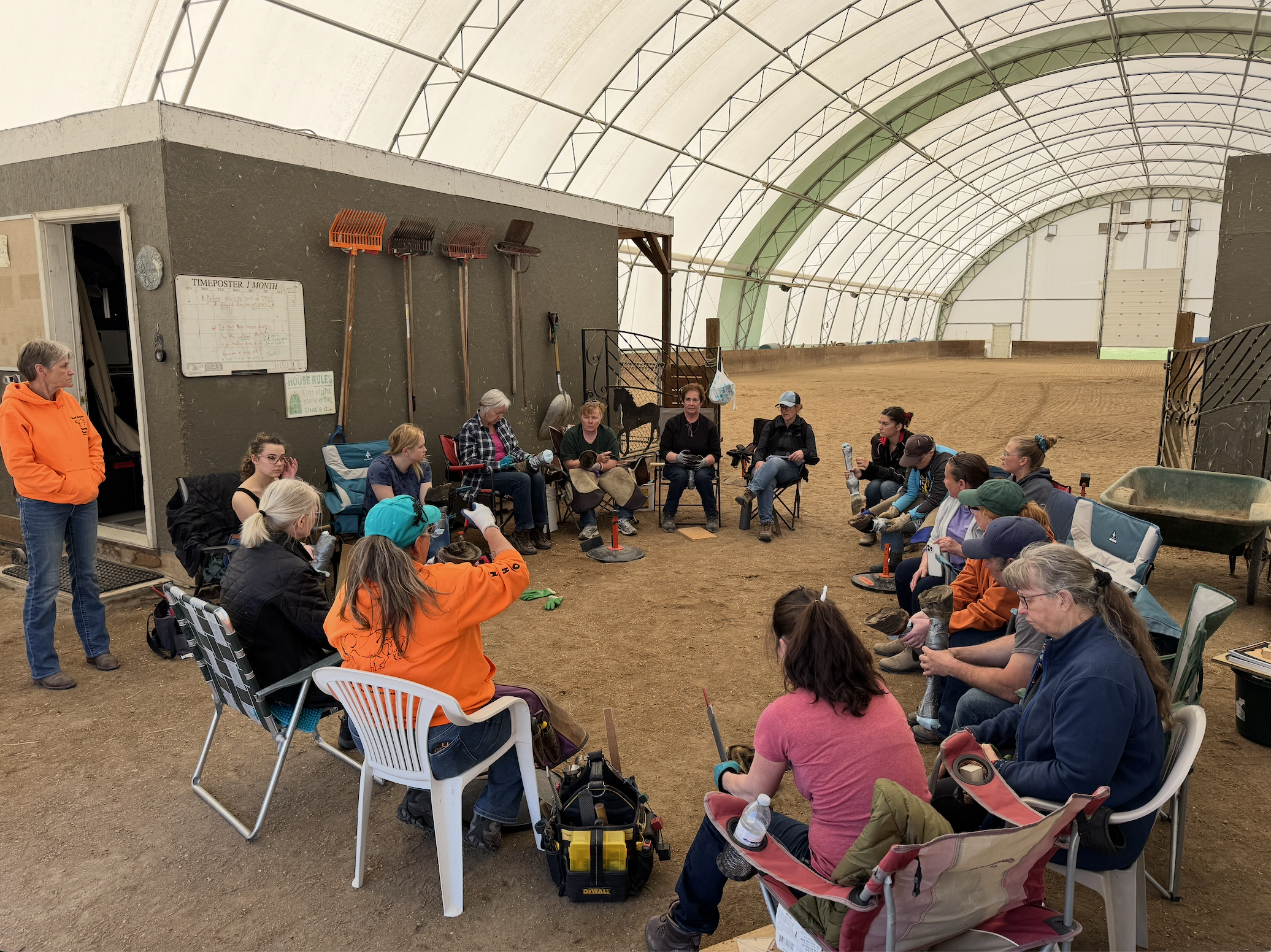 Group of people sitting in a circle inside an indoor horse riding arena, with some holding hoof cadaver and others listening. The arena has a dirt floor, a high curved ceiling, and a small building on the left with cleaning tools hanging on the wall.