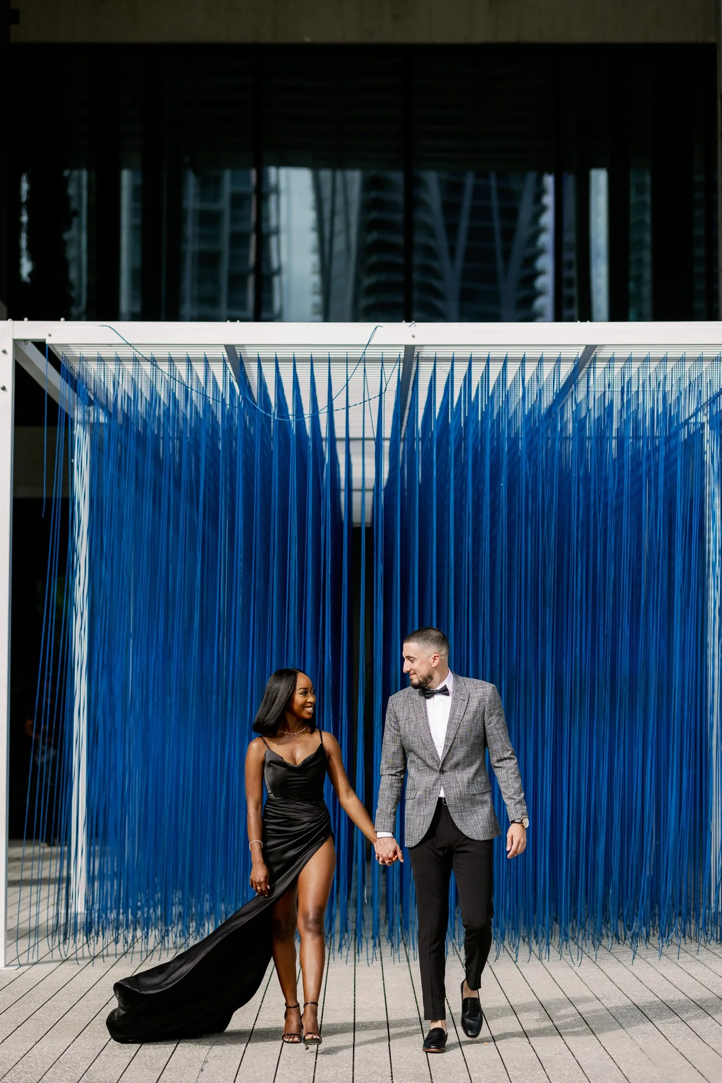 A stylishly dressed couple holding hands and walking in front of a contemporary art installation made of cascading blue strings, with the city skyline visible in the background.
